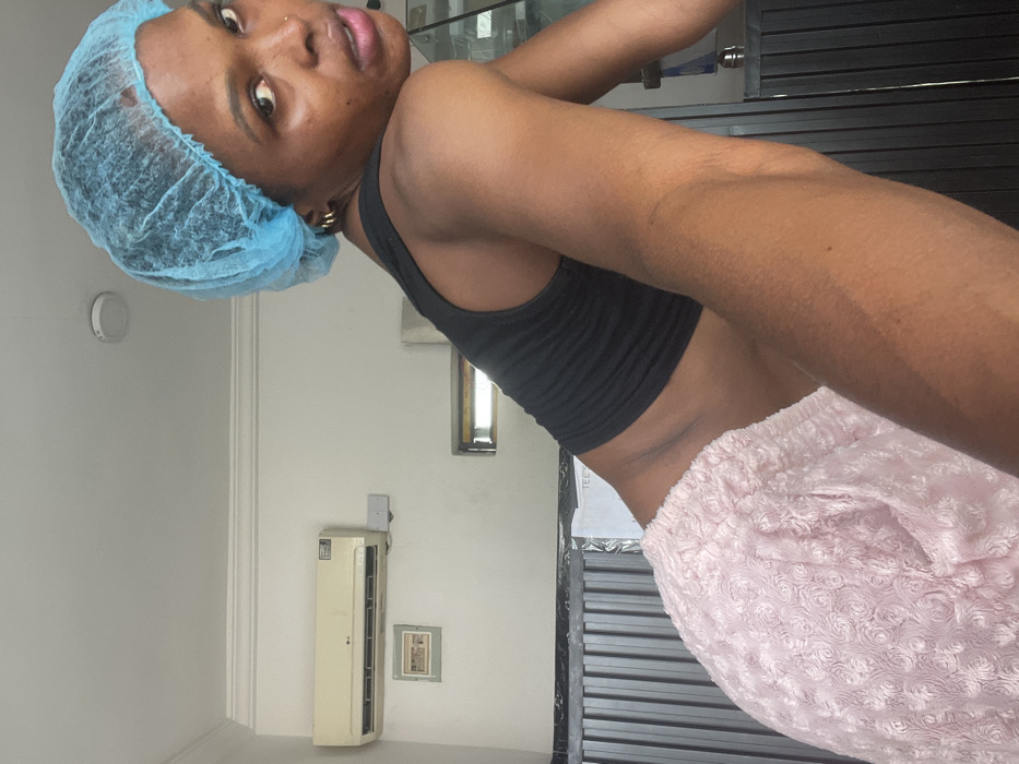 A woman wearing a blue disposable surgical cap, a black crop top, and fuzzy pink sweatpants poses indoors. Her dark skin is visible, and she wears a nose ring and gold hoop earrings. She looks towards the camera with a slight tilt of her head. Behind her is a white wall with an air conditioner unit and a small framed picture. A window and a closed black shutter are also visible in the background.