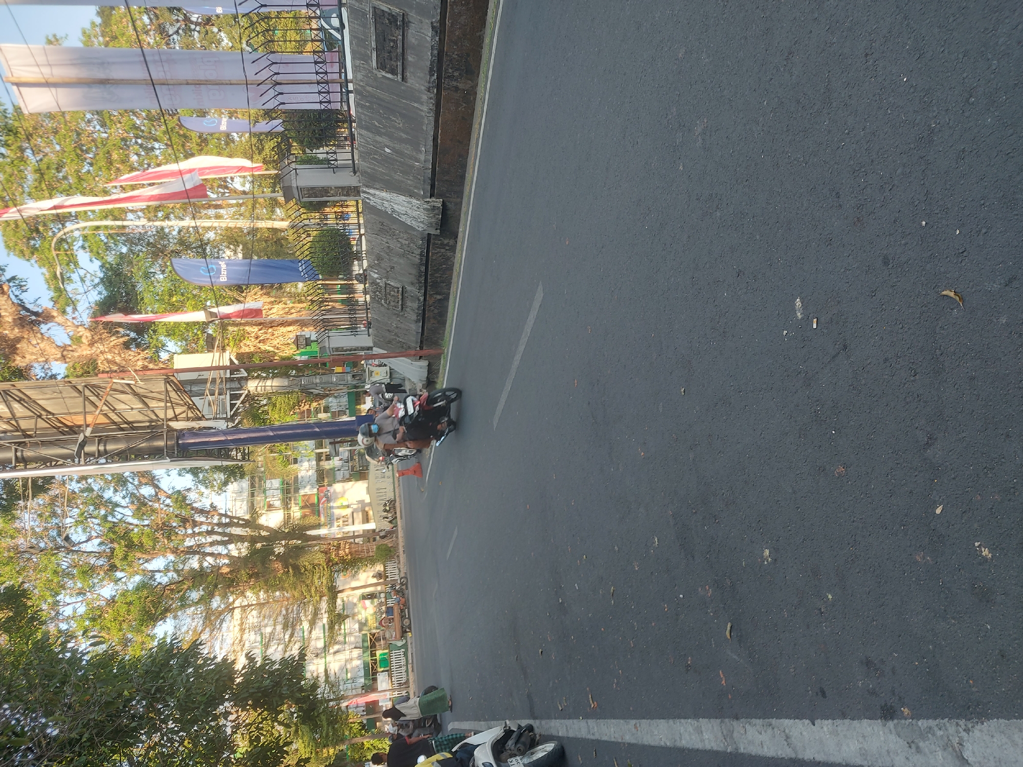 The image captures a street scene in Makassar, Indonesia, bathed in the warm light of either early morning or late afternoon. The perspective is from ground level, looking down a moderately busy street. In the foreground, the asphalt road is marked by a white line on the left side. Scattered on the road are dry leaves.
On the left side of the street, a white scooter is parked, with its front wheel pointing towards the viewer. A person in a green hijab and a light-colored shawl stands near the scooter. Further down the street, a few more parked motorcycles are visible.
Moving towards the center of the image, a person wearing a blue helmet and a mask is riding a black motorcycle, with a passenger behind them also wearing a helmet and mask. The motorcycle is leaning into a slight turn. In the background, there are several flags, including red and white Indonesian flags and blue flags with the "Biznet" logo. Behind the flags, a multi-story building with many windows is visible, partially obscured by tall trees with lush green foliage. A tall advertising billboard structure stands prominently near the center of the frame. A gray concrete barrier with a metal fence runs along the right side of the road, separating it from the sidewalk and building. The overall atmosphere suggests a typical urban environment with moderate traffic and daily activities.