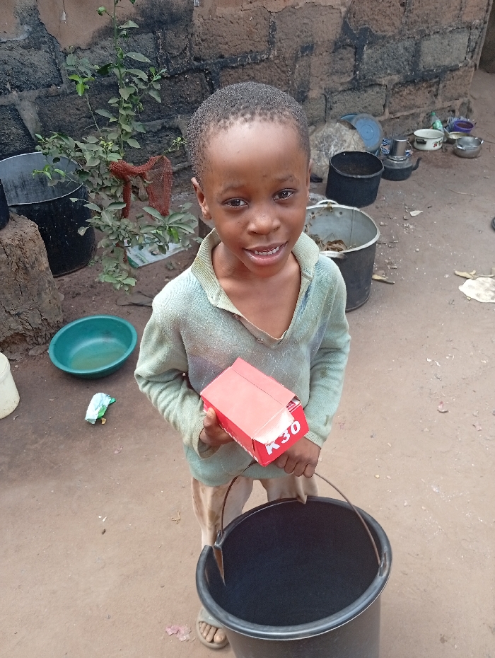A young boy stands outdoors holding a red box labeled "K30" in his left hand and a black bucket by its handle in his right hand. He's wearing a light-colored, collared shirt and light-colored pants. A green plant grows near a brick wall in the background. Various containers are visible in the background, including pots, buckets, and a green bowl. The ground is reddish-brown, suggesting a dirt or clay surface. The boy is looking directly at the camera with a slight smile.