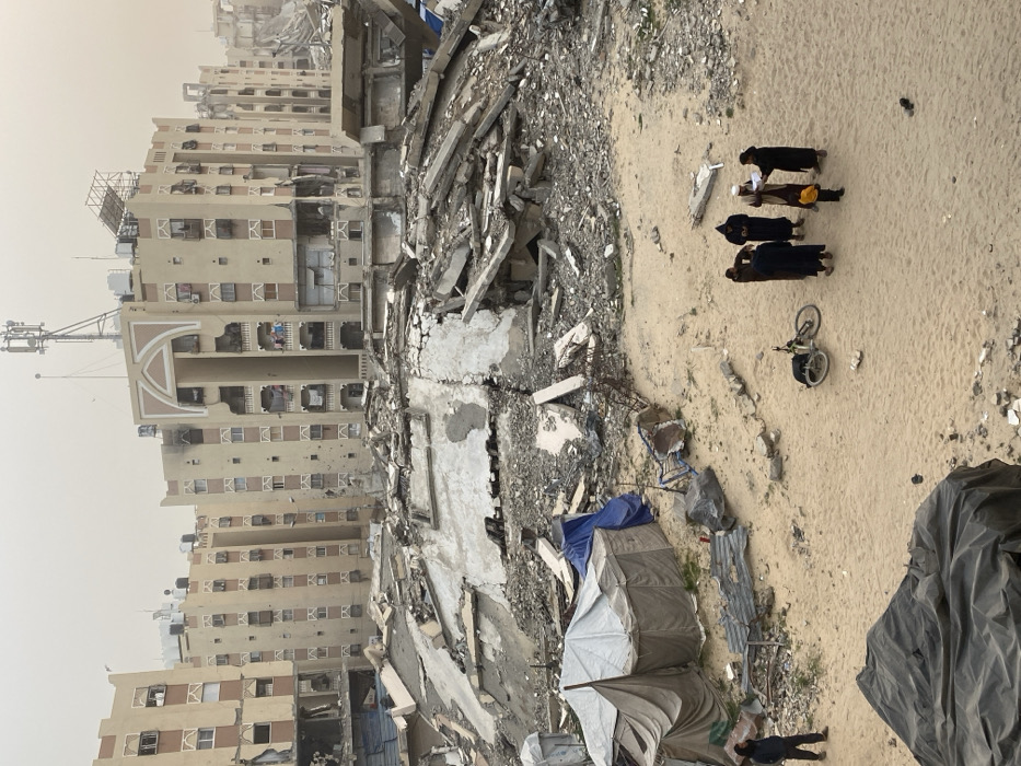 The image depicts a devastated urban landscape in Khan Yunis, Palestinian Territory, under an overcast or hazy sky. In the background, multiple multi-story residential buildings stand, many exhibiting severe structural damage, including partially collapsed sections, blown-out windows, and blackened facades indicating fire damage. Some buildings retain architectural details like arched entryways and balconies, where laundry is visible on less damaged sections. Communication antennas, satellite dishes, and water tanks are present on rooftops.
The midground is dominated by large, irregular piles of concrete rubble, twisted rebar, and debris from collapsed structures, forming a chaotic mound of building remnants. To the left of this rubble, several makeshift tents constructed from light-colored canvas and blue tarpaulins are erected on the sandy ground, suggesting temporary shelter. Scattered metallic debris and broken objects are visible near these tents.
The foreground consists of a relatively open, sandy ground area. On the right, a group of five individuals is gathered. Four adults, dressed in dark, long garments, some with head coverings, surround a smaller child in a yellow top. One adult holds and appears to be reading from a white sheet of paper. A bicycle lies on its side in the sand near this group. To the far left, a lone male figure in dark clothing and a head covering stands facing towards the right. A large, dark tarp or fabric bundle is partially visible in the bottom left corner. The overall scene conveys widespread destruction and makeshift living conditions.