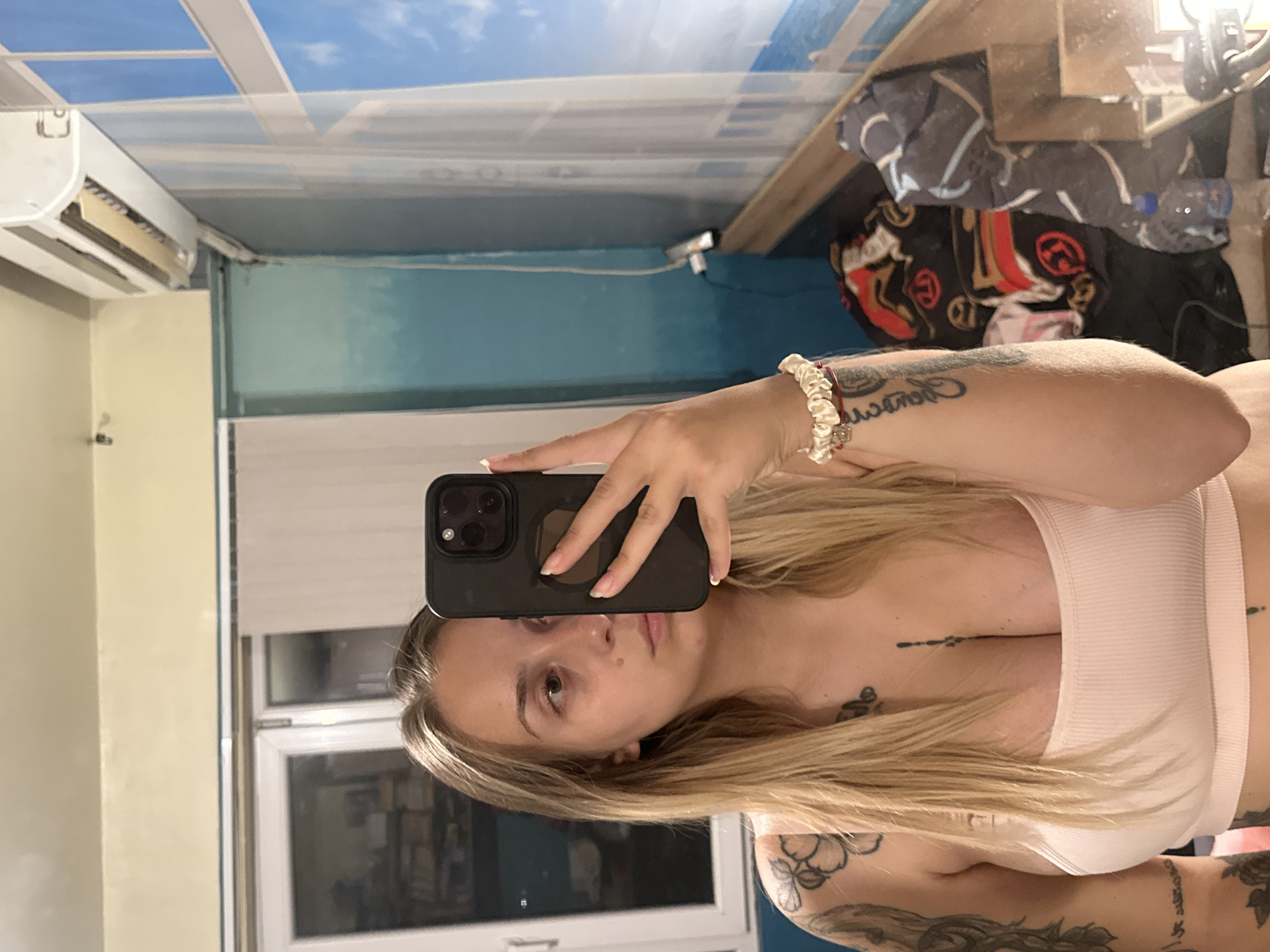 A young Caucasian female with blonde hair is taking a mirror selfie in what appears to be a bedroom. She is wearing a light pink ribbed tank top and has several tattoos on her arms and chest. She is holding a black smartphone in her right hand, partially obscuring her face. In the background, there is a window with a view of a blue sky and clouds, an air conditioning unit mounted high on the wall, and a cluttered area with blankets and a water bottle. The overall environment suggests a casual indoor setting. This image was not taken in the city of Plovdiv, Bulgaria.