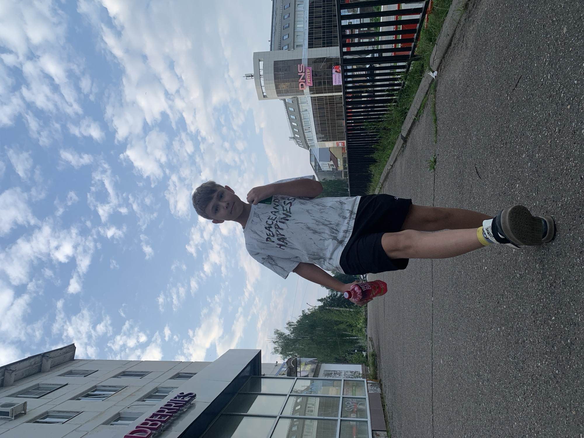 A young Caucasian male with short brown hair is walking towards the camera on a paved sidewalk in Chaikovsky, Russia. He is wearing a white t-shirt with a marble-like pattern and the words "PEACE AND HAPPINESS" printed on it, black shorts, yellow socks, and dark sneakers. In his right hand, he holds a red bottle of what appears to be a beverage. The sidewalk is lined with a black metal fence and overgrown grass on the right. To the left of the sidewalk, there is a white building with multiple windows and signage that reads "BERRIES". Further in the background, across the street, is a modern building with large glass windows and prominent signage displaying "DNS ГИПЕР". The sky is partly cloudy with scattered white clouds against a blue backdrop. The overall lighting suggests it is daytime.