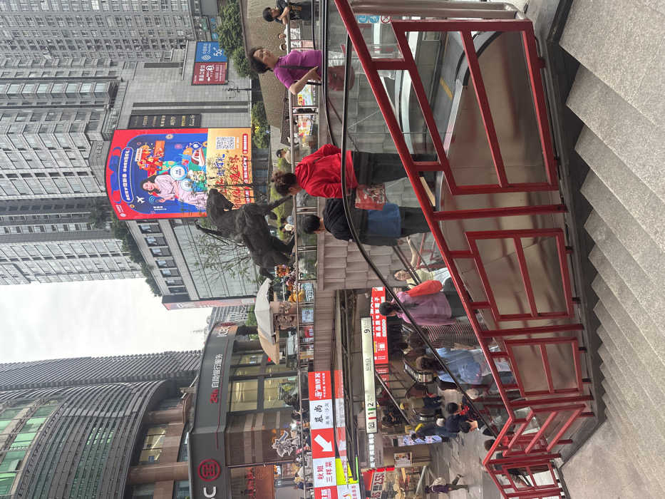 The image shows a scene in Chongqing, China (29.557, 106.572), featuring a red metal railing descending towards a set of stairs. The railing is attached to a structure that appears to be an escalator. The escalator is located in a bustling, urban area with a tall building in the background adorned with a billboard featuring a woman in a red dress. The woman in the red dress is a prominent subject, and her presence contributes to the urban ambiance. People of different ages and genders walk up and down the escalator, navigating the busy pedestrian area. The scene is a common occurrence in this bustling Chinese city, showcasing the vibrant and dynamic nature of life in Chongqing.
