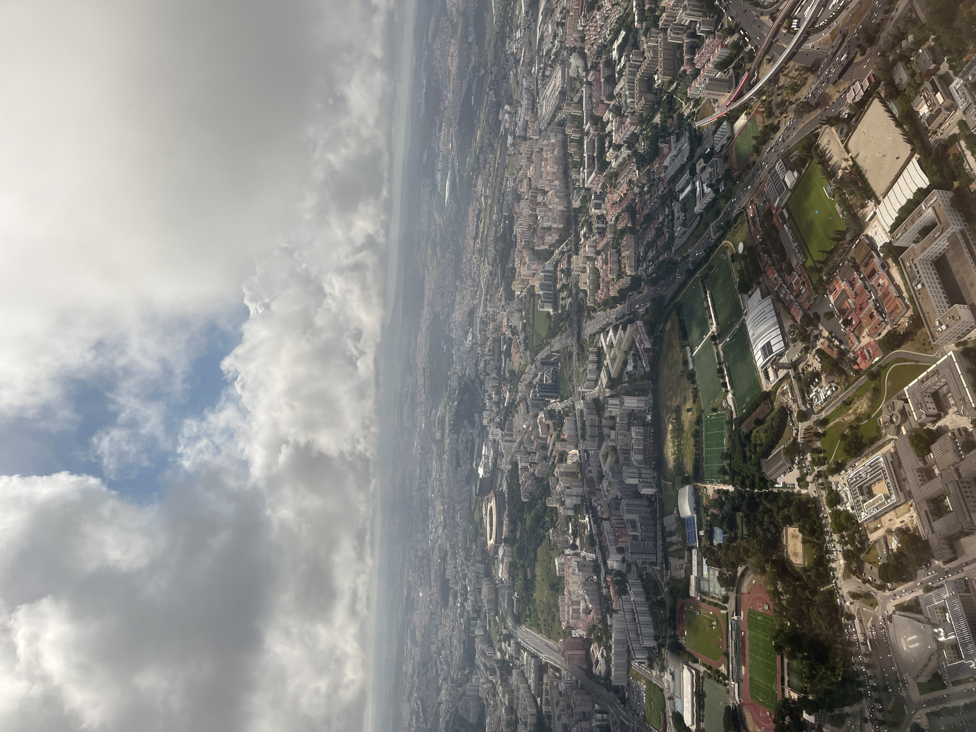 The image is an aerial view of Lisbon, Portugal. The top right shows buildings, roads, and green spaces, including athletic fields and tracks. In the middle is a stadium, surrounded by urban development. The left side is dominated by heavy cloud cover, giving the impression of looking down from an airplane. The city layout shows a dense urban environment with a mix of residential and commercial buildings. The weather appears overcast.
