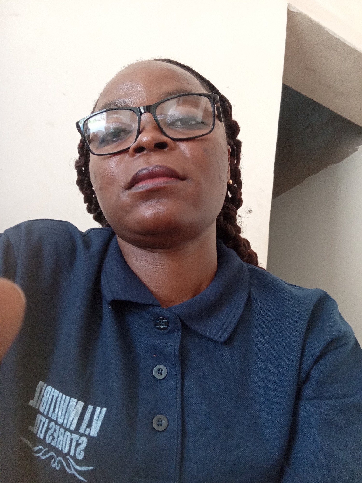 A waist-up, close-up, selfie-style photograph shows a dark-skinned woman with braids and glasses. She is wearing a navy blue polo shirt with a collar and three buttons. The shirt has white lettering on the left side of the chest, which appears to be a logo or text, but it is partially obscured and reversed due to it being a reflection from a mirror. The background is a plain, light-colored wall with a vertical shadow on the right side, suggesting an indoor environment with a light source from the left. The image was taken in Jalingo, Nigeria.