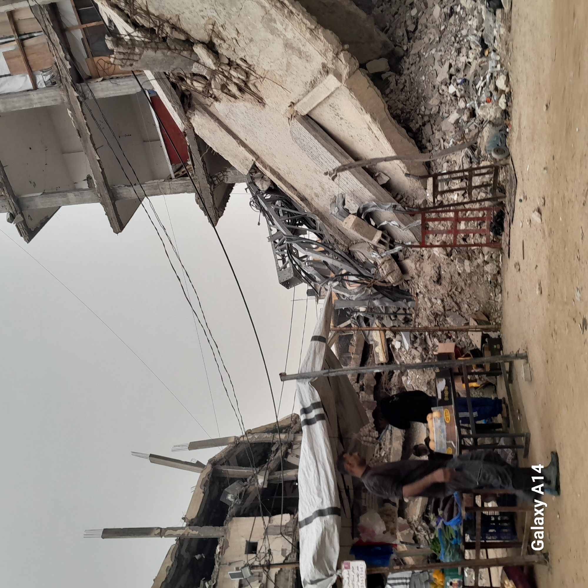 The image depicts a scene in what appears to be Khan Yunis, Palestinian Territory, judging by available context and details visible. A partially collapsed building dominates the background, with debris strewn about. Power lines are visible overhead. In the foreground, a man stands next to a makeshift structure, possibly a market stall. It holds various items, including what looks like packaged goods and textiles. The man seems to be looking or attending to items on the stall. The overall atmosphere suggests destruction and a struggling informal economy.
