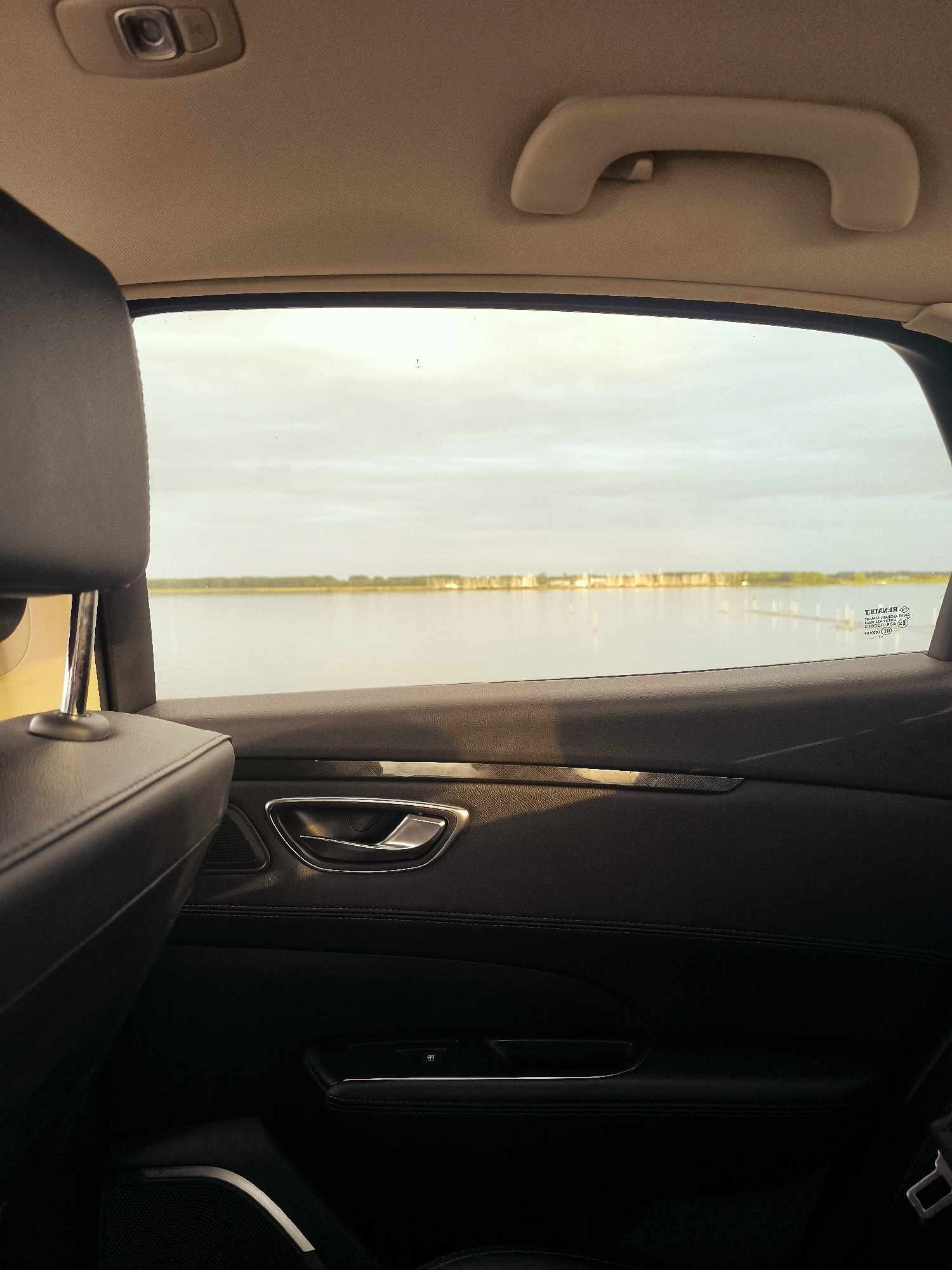 The image shows the interior of a car, looking out of the rear window. The view outside the window is of a calm body of water, likely a canal or lake, with a thin line of trees and buildings along the distant shore. The sky is overcast. Inside the car, the black leather interior of the door panel is visible, featuring a chrome door handle and a window control button. The headrest of the car seat is also visible in the foreground. The overall impression is of a quiet, perhaps early morning or late afternoon scene, viewed from a stationary vehicle. Based on the presence of the water and the architectural style of the buildings visible in the distance, this scene could be located in Lelystad, Netherlands, known for its proximity to water and modern architecture.