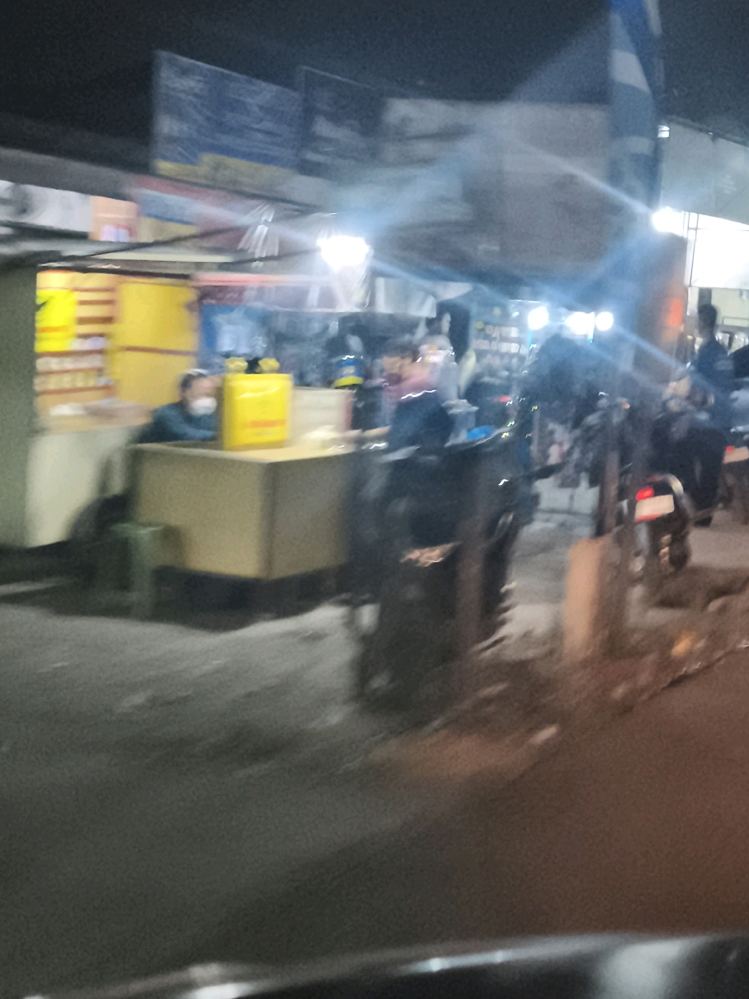 This image depicts a blurry, nighttime street scene in Palembang, Indonesia, possibly near a food stall or market. A person wearing a face mask is seated behind a counter. Various yellow boxes are visible on and around the counter. The background shows blurred signs and lights, suggesting a commercial area. Several motorcycles are parked or in motion, with one visible with its taillight illuminated. The overall impression is that of a busy, urban environment after dark.