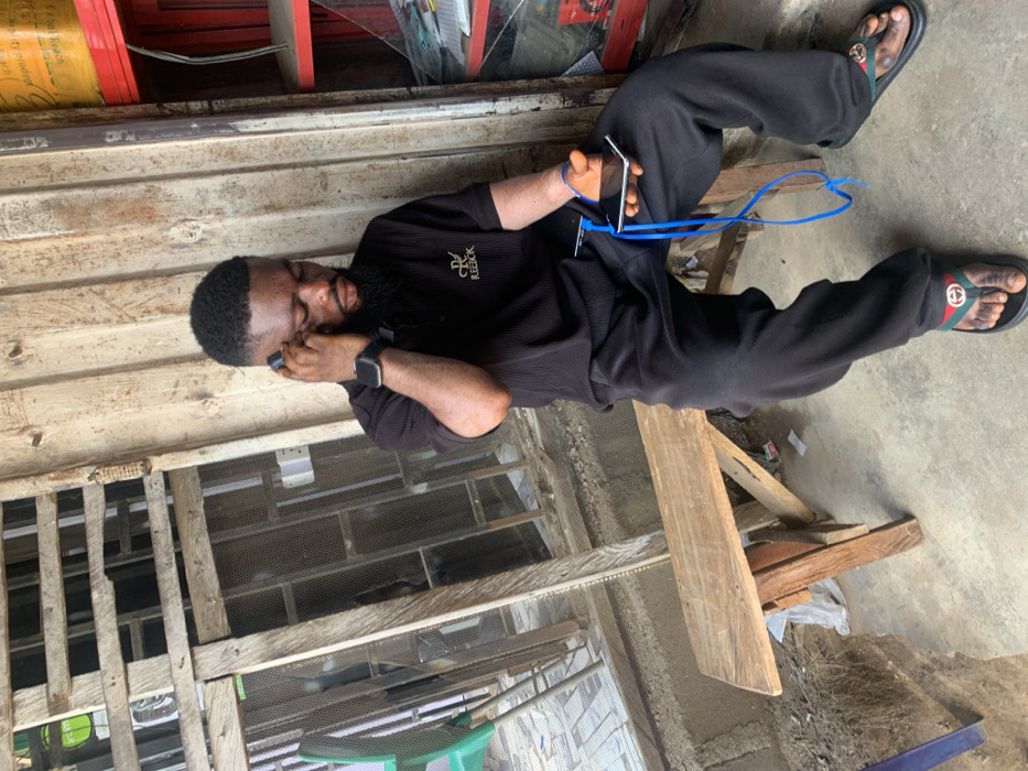 This image depicts a male subject seated on a rustic wooden bench within an urban environment in Okene, Nigeria. The individual, of dark complexion with a short beard and dark hair, is wearing a black short-sleeved collared shirt with a gold "RL REEBOK" logo on the left chest, black trousers, and black slide sandals with green and red striped accents. A black smartwatch is visible on his left wrist.
He is engaged with two mobile phones: one black smartphone is held to his left ear, suggesting an ongoing call or audio interaction, while a second silver-backed smartphone is held in his right hand. A blue cable, likely a charging cable, is connected to the phone in his right hand and extends downwards. His gaze is directed downwards and to his right.
The immediate background to the left features a weathered wooden structure, possibly a window frame or a security grill, with mesh visible and hints of greenery beyond. To the right, vertical wooden planks form a wall, behind which red and yellow shelves hold various items, including a yellow cylindrical container with visible text "Product of Nigeria, Ajao Beach," and a glass display case below. The ground is a concrete slab, appearing worn and somewhat uneven, with dirt and debris visible near the base of the wooden structures. The overall scene suggests an informal commercial setting, such as a shop entrance or a market stall.