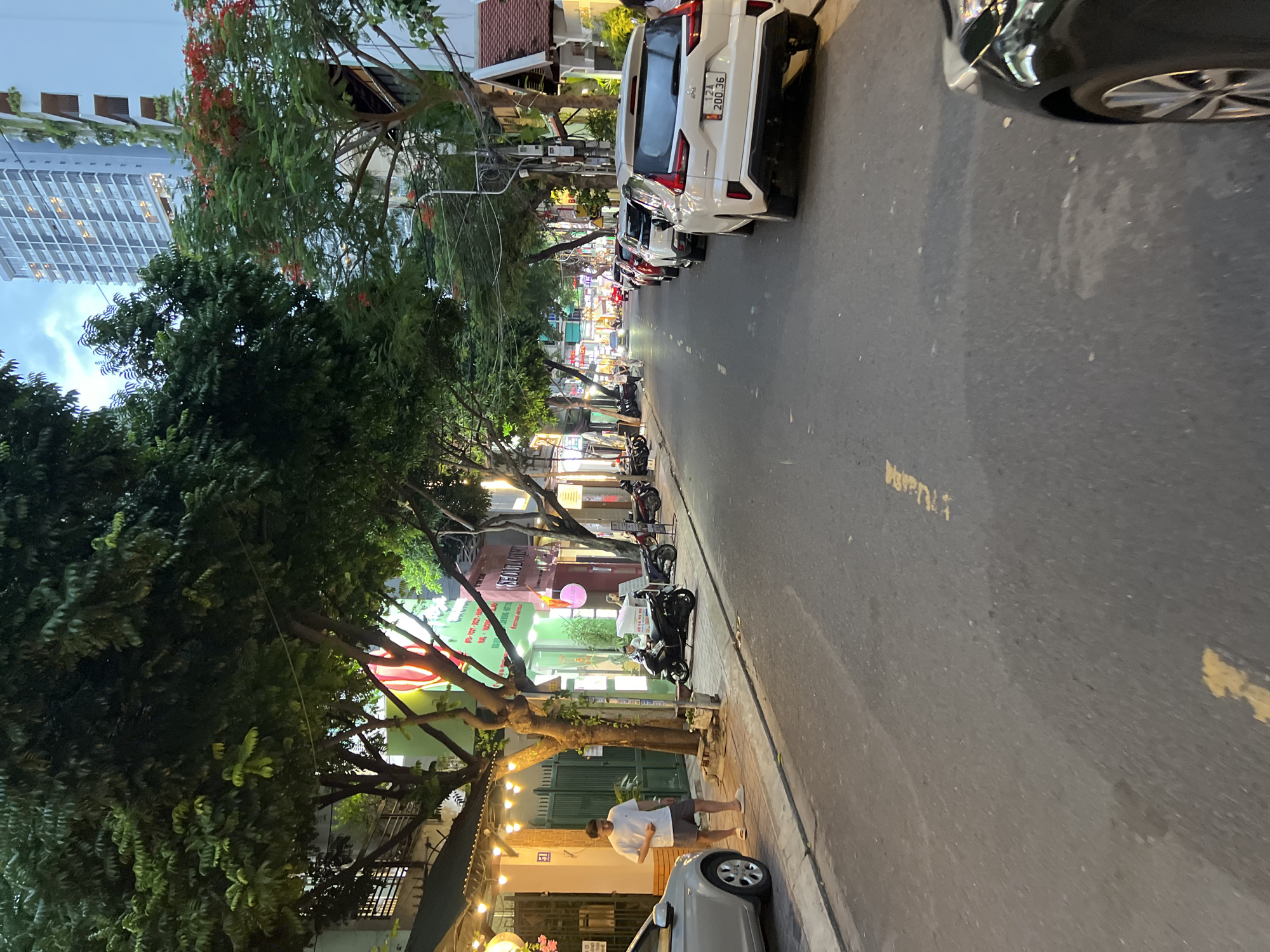 The image depicts a street scene in Nha Trang, Vietnam, during twilight or early evening. The street is lined with trees casting shadows, and buildings of varying heights, including a modern high-rise. Several cars and motorcycles are parked along the street and on the sidewalk. A man in a white t-shirt and dark shorts stands on the sidewalk near a parked silver car. A white SUV is prominently parked on the right side of the road, with its license plate visible. Yellow markings are present on the asphalt road. The lighting suggests artificial streetlights and lights from businesses are illuminated. The overall impression is a busy urban street in Vietnam.