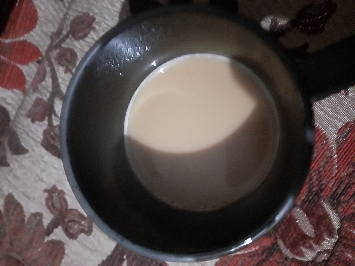 The image captures a top-down view of a dark-colored mug or small saucepan filled with a light brown, creamy liquid, most likely tea or coffee with milk. The container appears to be made of a dark material, possibly metal, and has a subtle handle visible extending to the right. The surface of the liquid is smooth and shows a distinct curved reflection, indicating a light source from above and slightly to the left. The container is placed on a patterned fabric surface, which serves as the background. This fabric features repeating floral or leaf motifs in shades of maroon or deep red, intermingled with lighter brown or beige tones, against a lighter, possibly off-white or light brown base. The overall lighting suggests an indoor setting. No people or text are visible in the image.