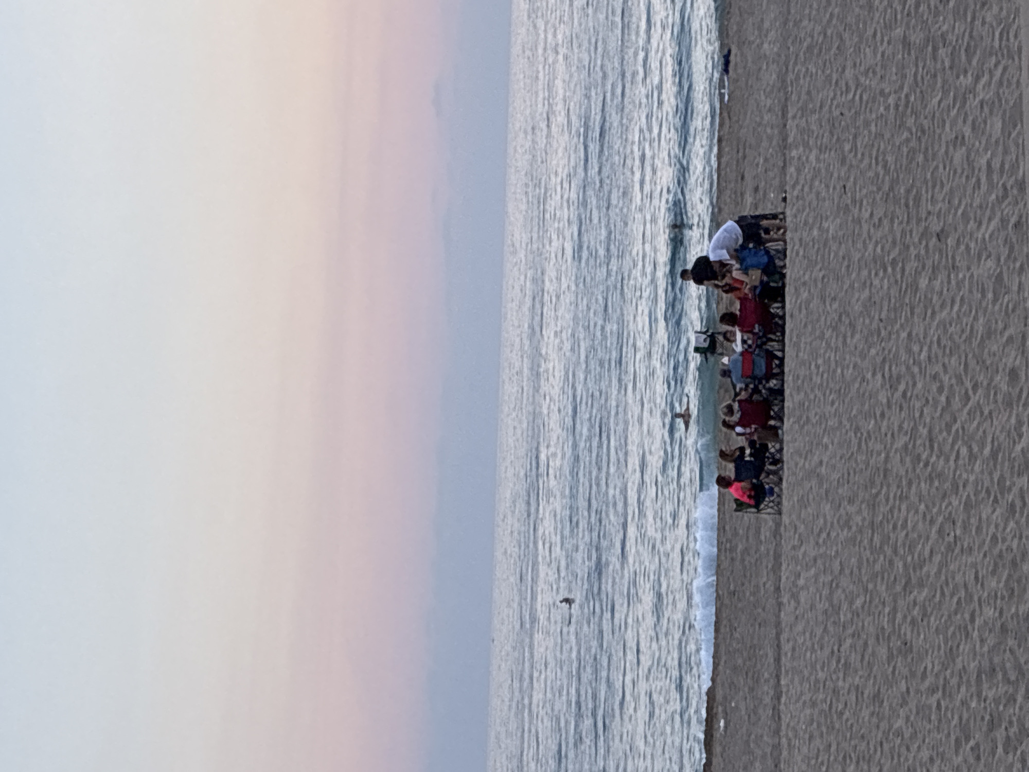 A group of people are gathered on a sandy beach, facing the ocean. The sky is a gradient of pale blue to pink, indicating either sunrise or sunset. Several individuals are seated in portable chairs around a low table, some appear to be talking or interacting. One person, wearing a white t-shirt and dark shorts, stands near the seated group, possibly engaged in conversation or handing something to another person. In the ocean, several people are visible, some swimming or wading in the water, and at least two individuals are on surfboards further out. The waves are moderate, with white foam crashing on the shore. The sand appears textured, with visible footprints or patterns. The overall scene suggests a relaxed gathering on the beach. This scene is consistent with the beaches in the city of Alanya, Turkey, known for its coastal location on the Mediterranean Sea.