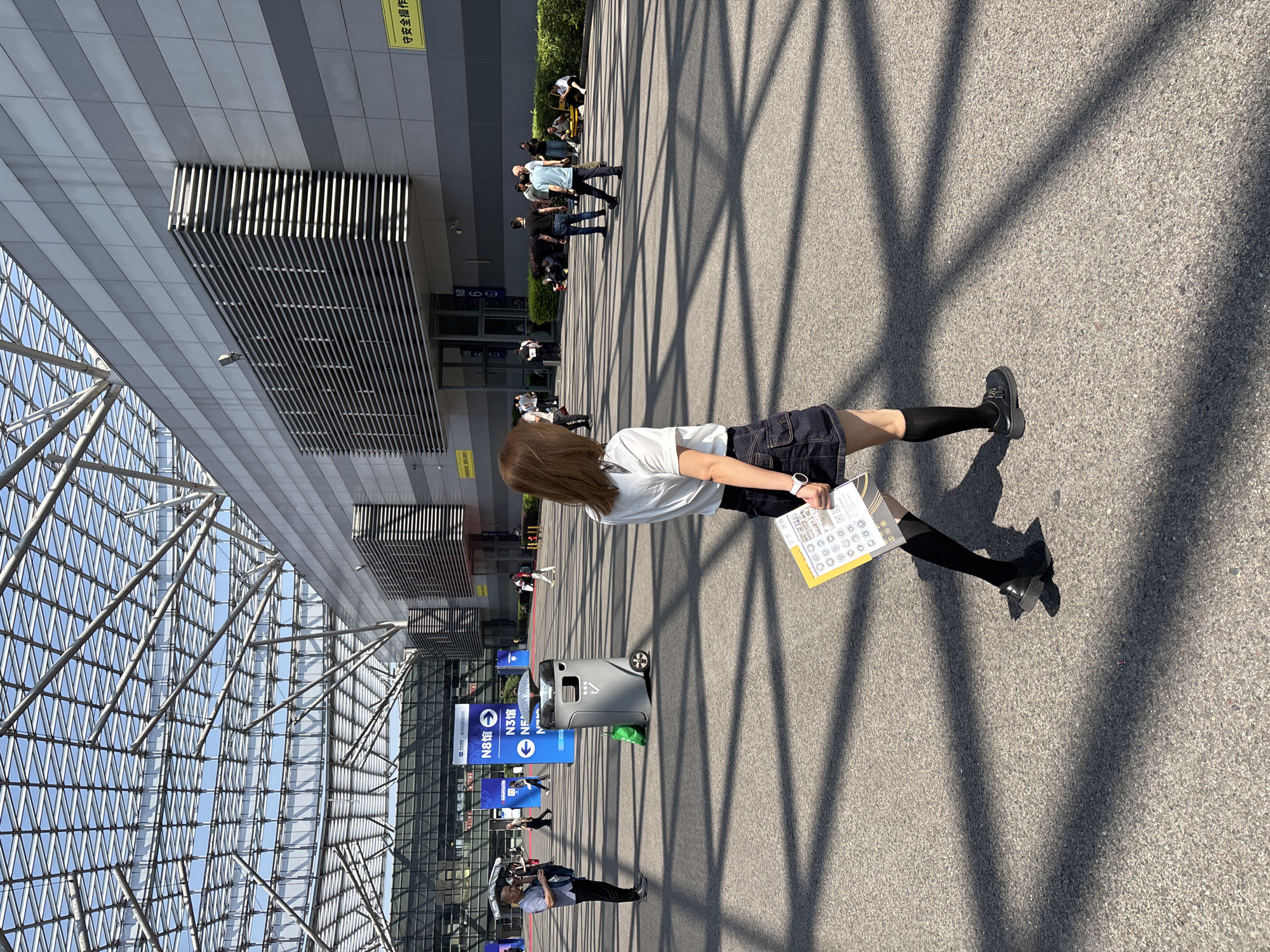 A woman in a white shirt, dark shorts, black socks, and black shoes stands on a concrete surface with shadows from a structural framework. She holds a yellow document with circular graphics. The location is outside a large building with a glass roof structure and metal facade. Blue signs are visible with directional information. Other people are present in the background. This scene is located in the city of Liangjiang, China.