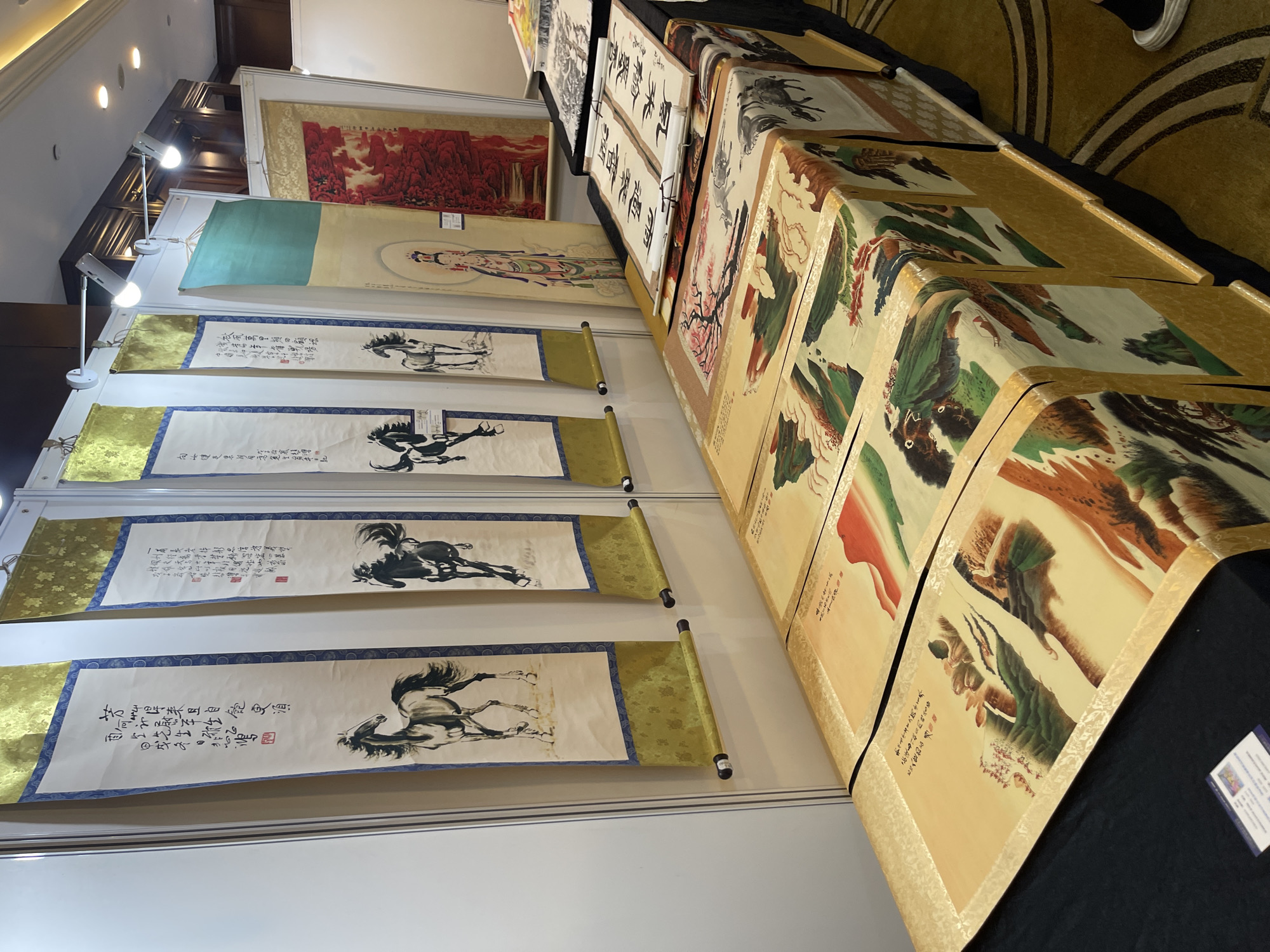 The image depicts a display of traditional Chinese scroll paintings, likely at an art exhibition or market. The scrolls are hung vertically and are presented in a row, suggesting a curated collection. The subjects of the paintings vary, with visible depictions of horses in dynamic poses, possibly in mid-gallop, and landscapes featuring mountains and foliage. One scroll appears to feature a figure, possibly a deity or a serene individual, in a flowing robe. The background suggests an indoor setting with spotlights illuminating the artwork. The scrolls themselves are rendered on what appears to be rice paper, some with ornate golden borders and dark rollers at the top and bottom. The overall presentation is neat and organized, with the scrolls unfurled to showcase the artwork. A small card with text and an image is visible on the bottom right, likely providing information about the artwork or artist. The setting appears to be well-lit, indicating it's likely daytime or a well-lit indoor venue. The presence of multiple scrolls and the way they are displayed points to an event focused on the appreciation or sale of Chinese art. The visible text, though partially legible, is in Chinese characters, common for such artworks.