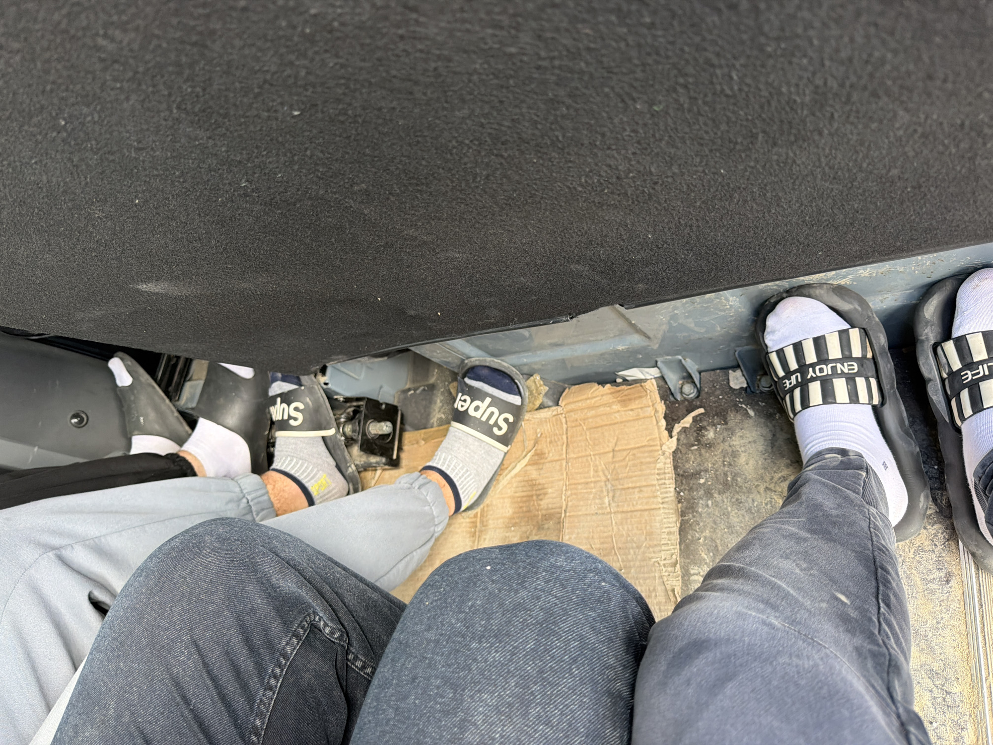 The image shows the feet of three individuals inside what appears to be a vehicle. The flooring of the vehicle is covered with torn cardboard and some metal components are visible. One person is wearing white socks and black slides with white stripes and the words "ENJOY LIFE" printed on them. Another person is wearing grey socks with the word "Super" printed on black slides. A third person is wearing white socks and black slides. The interior of the vehicle is dark and appears to be utilitarian. The scene is a close-up, low-angle shot focusing on the feet and lower legs of the individuals. No specific details about the city of Janowice, Poland, are discernible from the image.
