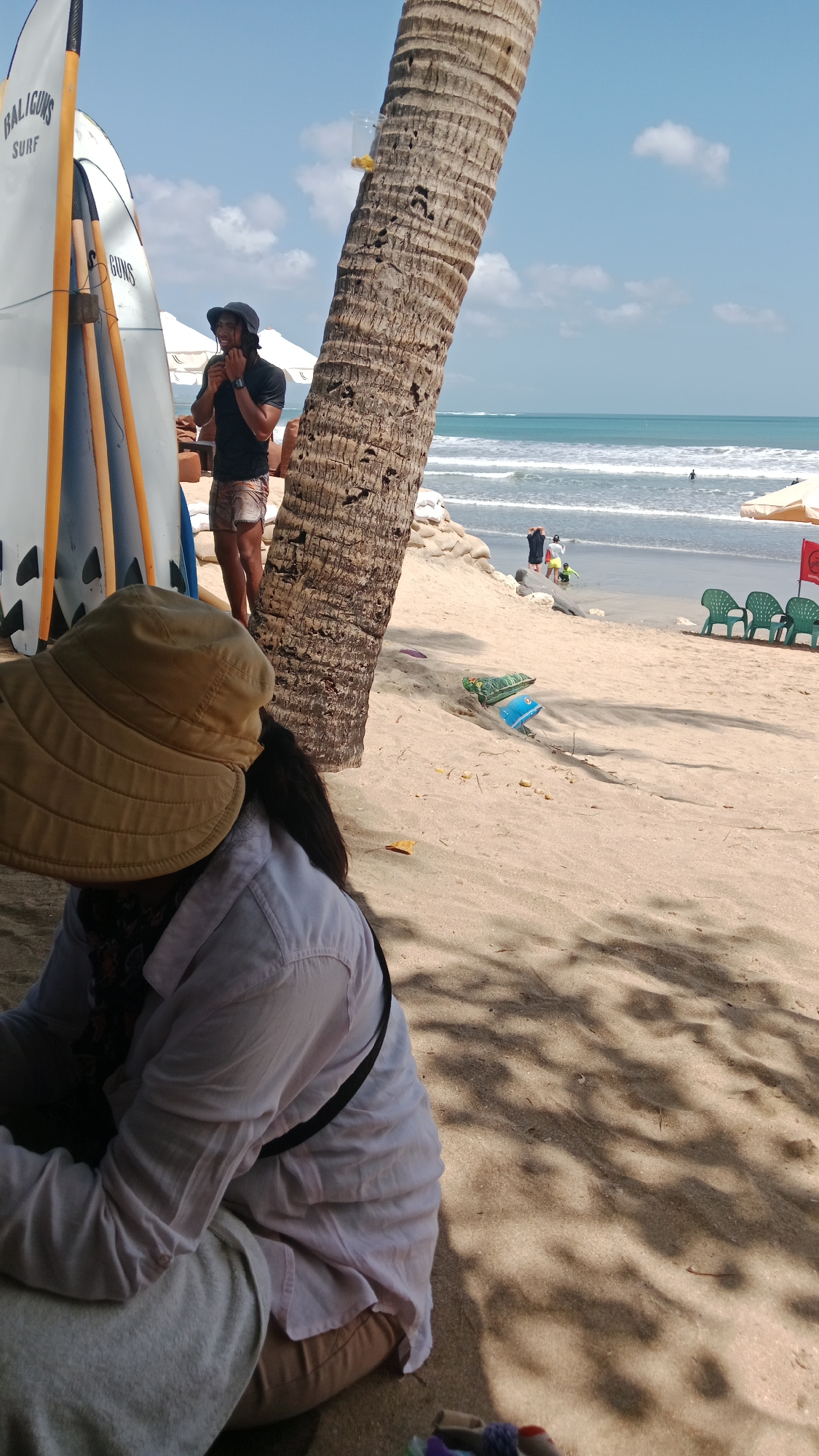 The image depicts a sunny beach scene in Kuta, Indonesia, under a clear blue sky with scattered clouds. In the foreground, a person wearing a tan sun hat and a light-colored long-sleeved shirt is seated on the sand, with their back to the camera. To the left, three surfboards stand upright, one white with orange fins and the others mostly obscured. A man in a dark t-shirt and patterned shorts, wearing a dark hat, stands near the surfboards, looking down and touching his face. A large palm tree trunk dominates the mid-ground, its rough bark textured by the sunlight. Beyond the palm tree, the ocean stretches to the horizon, with gentle waves breaking on the shore. Several beach chairs are visible on the sand, along with some scattered items. In the shallow water, a few individuals are present. The overall impression is a relaxed beach environment.