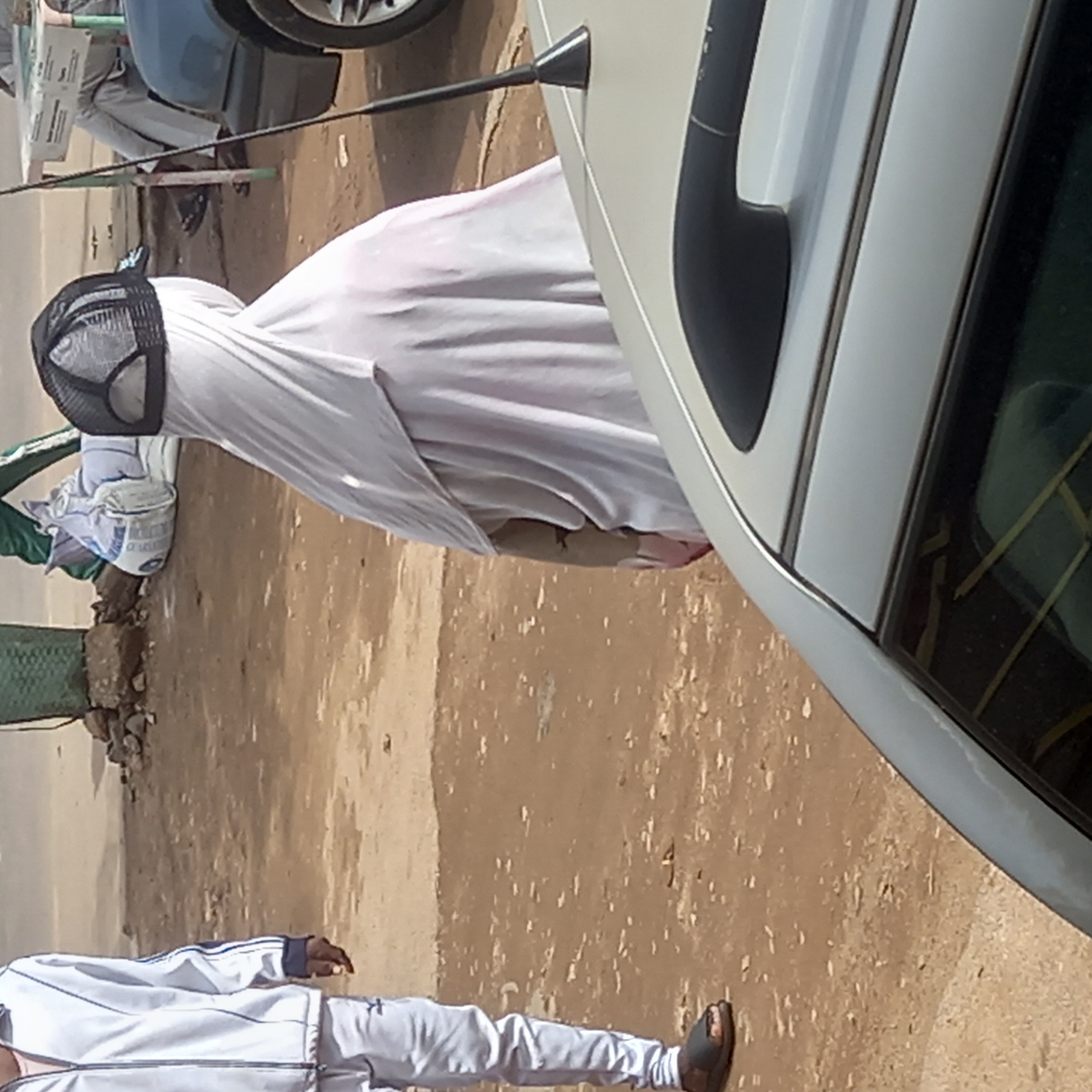 The image depicts a scene in what appears to be a public area in Dadi Riba, Nigeria. A person is fully covered in a white sheet, with only a black mesh cap visible on their head. They stand next to a parked white vehicle, with a portion of the car door and handle visible. In the lower foreground, the legs and torso of another person wearing a white tracksuit with blue stripes are visible. The ground appears to be a mix of dirt and paved surface. In the background, there are hints of greenery and other parked vehicles.