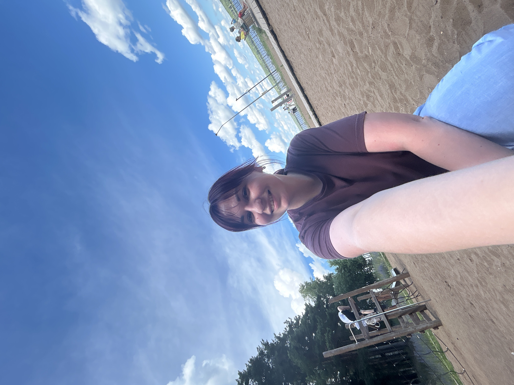 A young woman with dark hair and bangs smiles at the camera, holding out her arm to take a selfie. She is wearing a dark purple t-shirt and light blue jeans. She is sitting on a sandy beach, with a playground structure to her left and a wooden boardwalk extending into the distance to her right. Beyond the boardwalk is a body of water and trees. The sky is blue with scattered white clouds. The location appears to be a public park or recreational area.