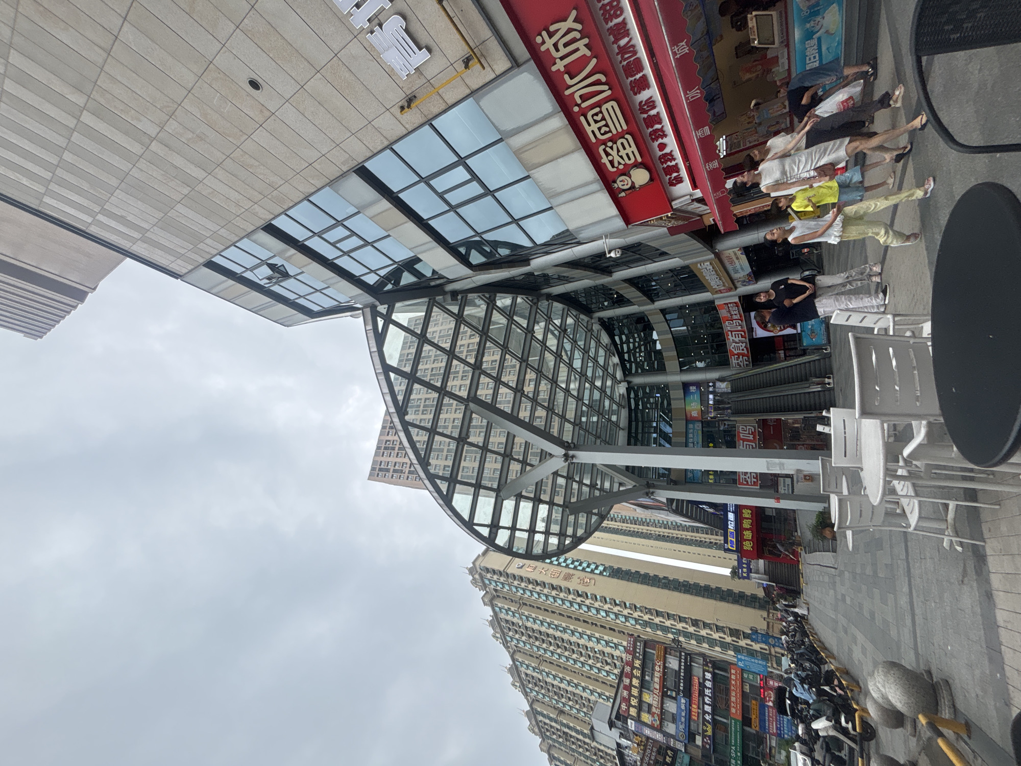 The image depicts a street scene in Nanhai District, China, on an overcast day. Dominating the right side of the frame is a modern building with a beige facade and large glass windows. A red awning with Chinese characters, reading "蜜雪冰城" (Mì Xuě Bīng Chéng, meaning Mixue Bingcheng, a popular ice cream and tea chain), extends across the lower portion of this building. Several people are visible walking along the sidewalk in front of the building. To the left, a large glass-covered structure with a metal frame shelters an escalator. Further to the left, a very tall, light-colored apartment building with numerous windows stretches into the sky. In the foreground, several white tables and chairs are set up on the paved walkway, suggesting an outdoor seating area for a cafe or restaurant. The ground is paved with grey tiles. In the mid-ground, a row of parked motorcycles and scooters lines the side of the street. The sky is filled with heavy, grey clouds.