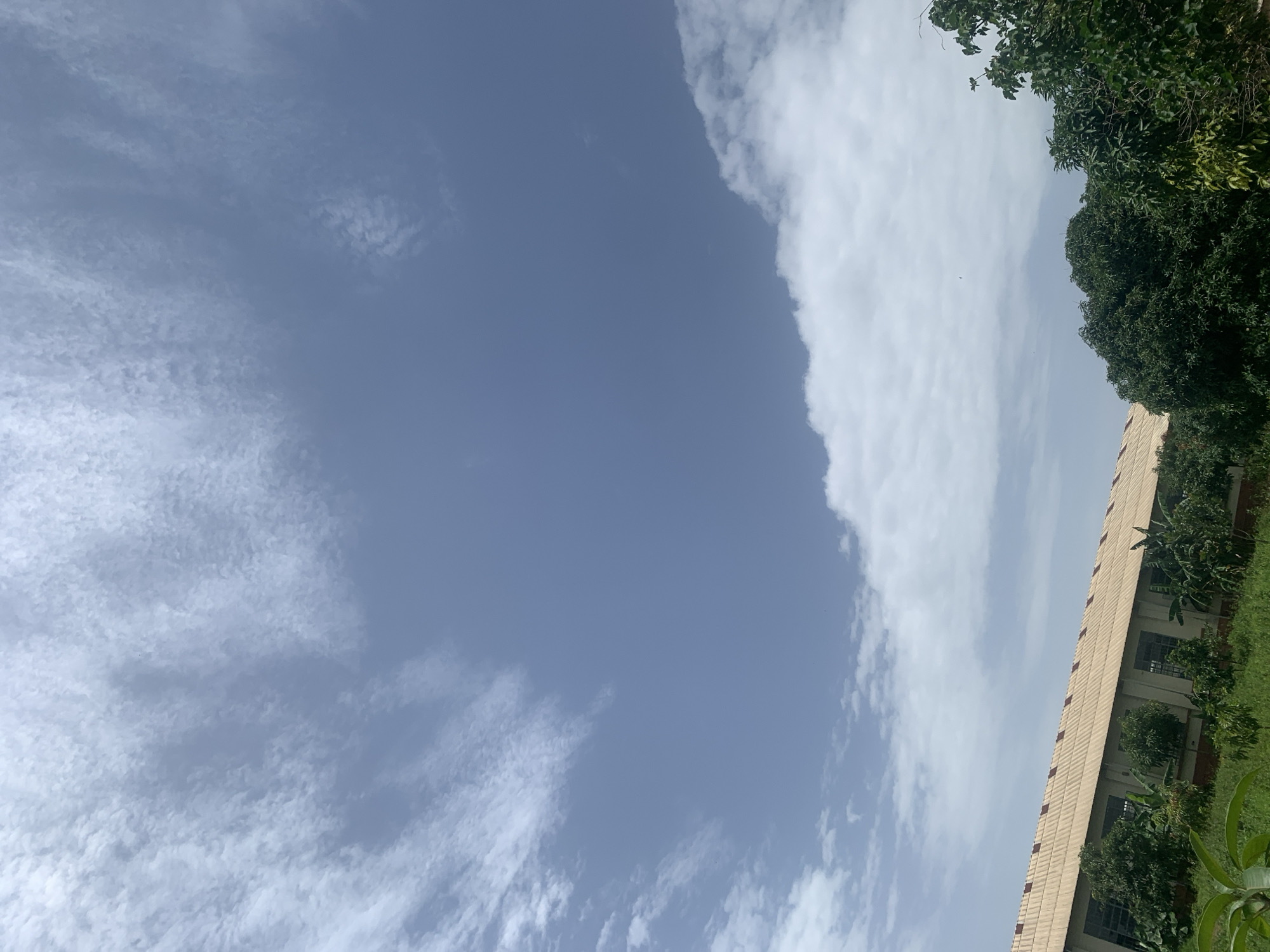 The image depicts a clear, bright sky with wispy white clouds. In the foreground, a long, low-rise building with a corrugated metal roof is visible. The building appears to be a warehouse or industrial facility, with numerous windows lining its facade. Surrounding the building is lush green vegetation, including trees and smaller plants. The overall scene suggests an outdoor setting in a semi-rural or industrial area of Kuru Karama, Nigeria. No people are present in the image.