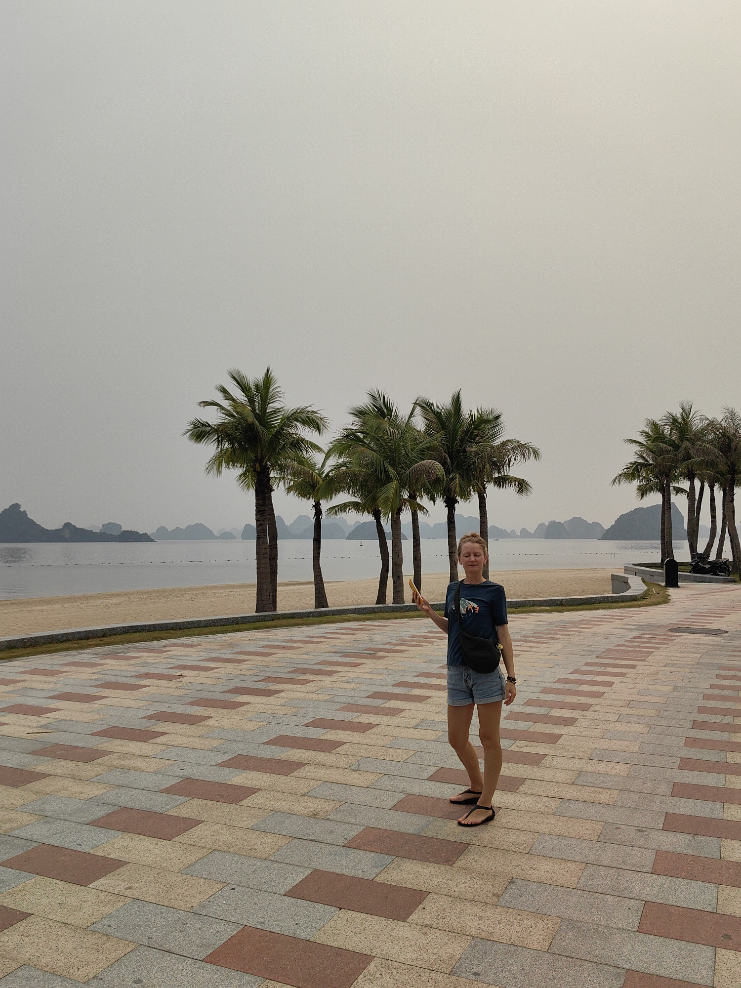 The image features a woman standing on a patterned paved walkway alongside a sandy beach and bay, characteristic of Ha Long Ward, Vietnam. The woman, positioned slightly left of center, faces the camera with her eyes closed or squinting and a gentle smile, holding a yellow smartphone in her right hand. She wears a dark blue t-shirt with a graphic design, light denim shorts, and black flip-flops, with a black fanny pack worn across her chest.
Behind her, several tall palm trees line the edge of the sandy beach, which borders a calm body of water. In the distance, numerous iconic karst islands or limestone formations rise from the bay, partially obscured by an overcast and hazy sky, which is a uniform light grey. A line of buoys or markers stretches across the water.
The weather appears hazy and diffused, creating soft, muted lighting. The paved walkway beneath her feet is adorned with a mosaic pattern of rectangular tiles in various shades of red, beige, and grey. The overall scene conveys a tranquil, relaxed atmosphere in a coastal setting during daytime, despite the overcast conditions. No explicit text is visible.