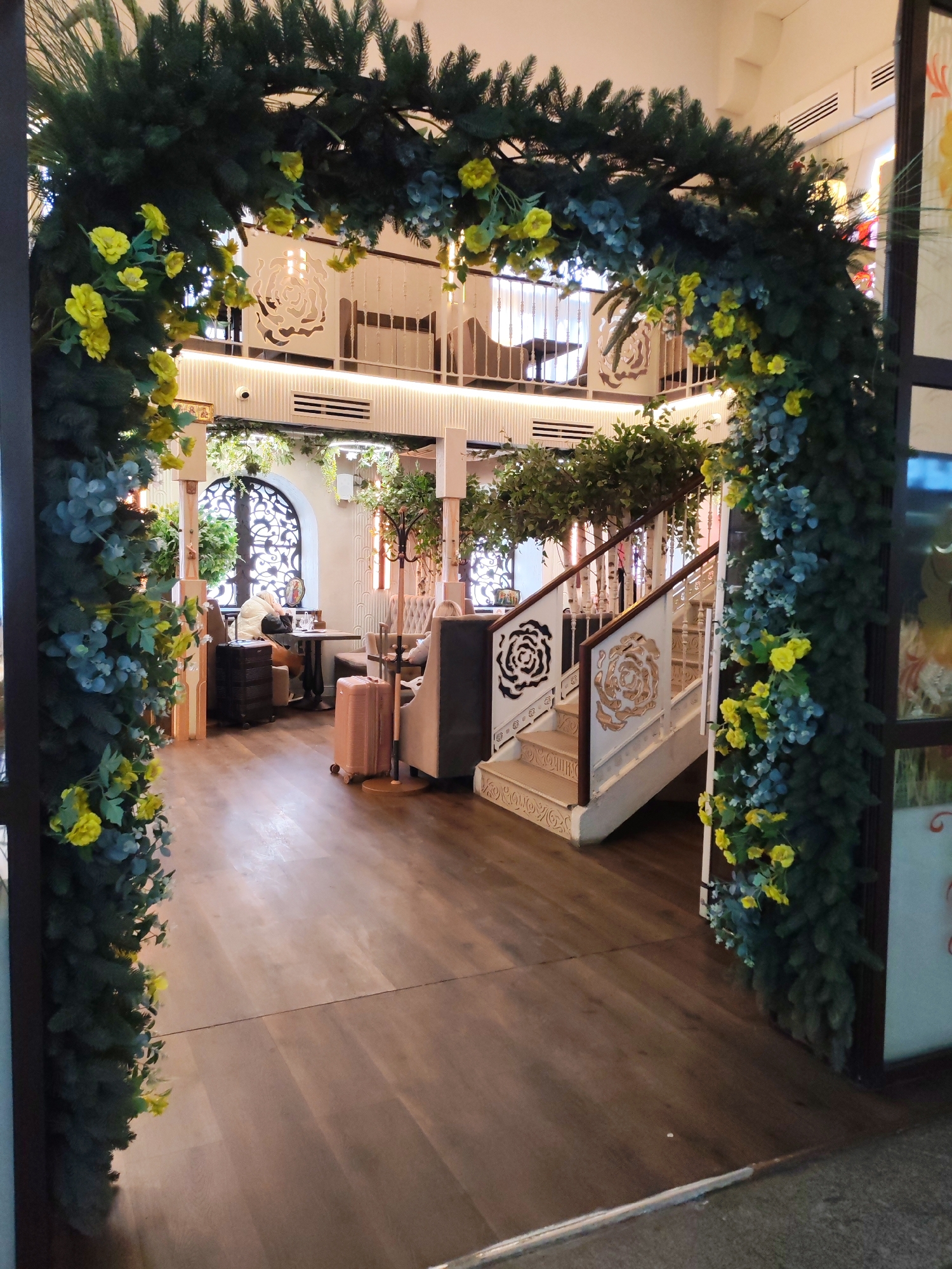 The image displays the elegant interior of an establishment, likely a restaurant, cafe, or hotel lobby, located in Moscow, Russia. A large archway, adorned with lush artificial evergreen foliage, bright yellow flowers, and light blue-green eucalyptus-like leaves, frames the entrance in the foreground.
Beyond the arch, the space reveals a two-story layout with warm wooden plank flooring and light-colored walls, some featuring vertical fluting. A prominent white staircase with wooden handrails ascends to the upper level. The staircase's side panels are decorated with large, stylized rose motifs, one in black and another in gold/bronze, a design echoed in white cutouts or reliefs on the upper walls.
Several patrons are seated at tables. On the left, a person is seen at a table next to a large arched window with intricate dark grillwork. Beside their table are two suitcases: a stack of black luggage and a single light pink/rose gold suitcase, suggesting travelers. Another person is partially visible seated on the right.
The venue features a strong botanical theme, with abundant hanging greenery and potted plants throughout the space, contributing to a fresh, garden-like ambiance. Lighting is provided by recessed ceiling lights, creating a bright and inviting atmosphere. A wooden coat rack stands near the staircase. No discernible text is visible within the image.