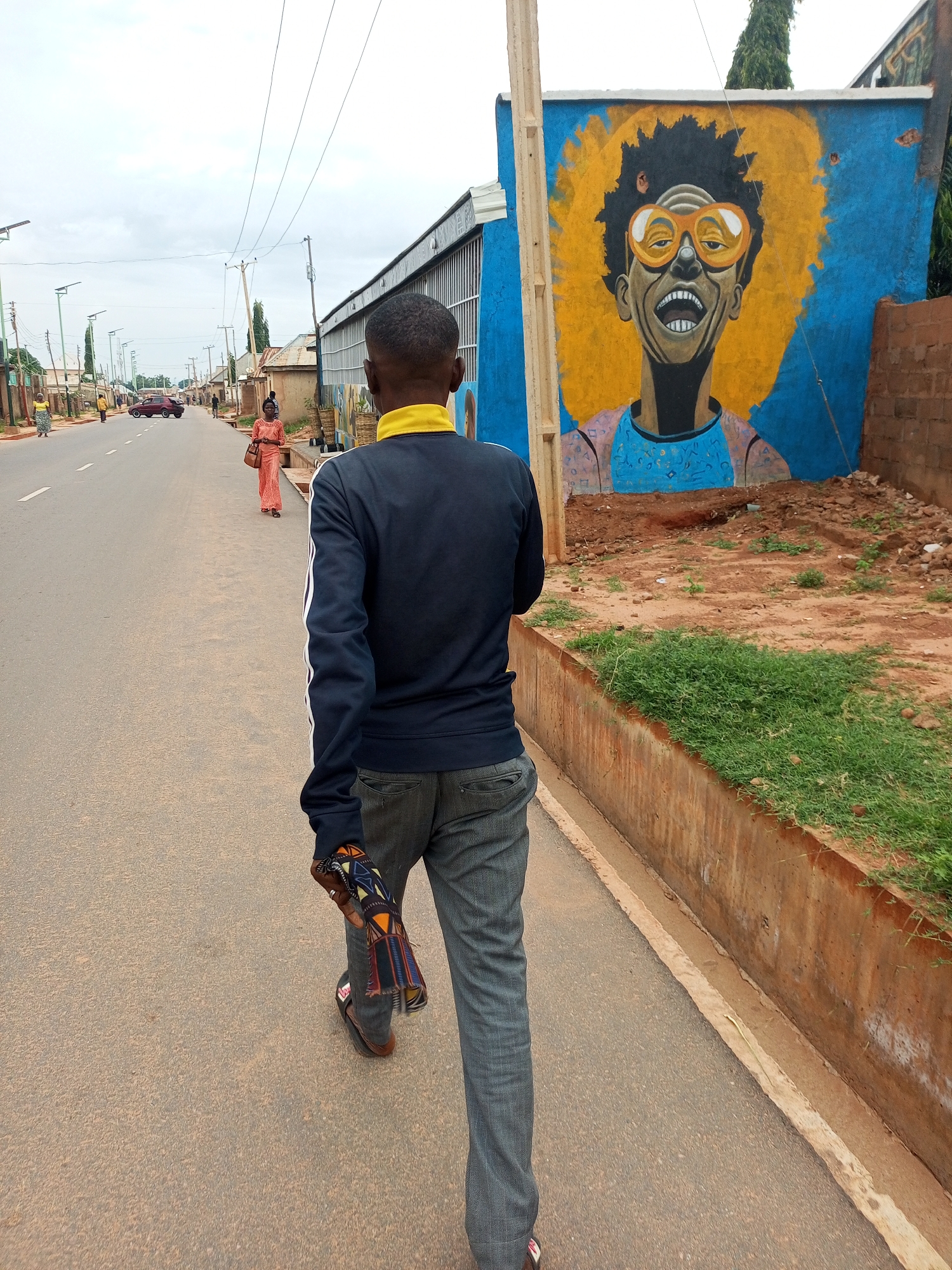 The image depicts a street scene in Tempure, Nigeria. In the foreground, a person walks away from the camera, wearing a dark blue jacket with yellow trim and grey pants. The person is holding a folded, patterned fabric in their right hand. To the right of the street, a vibrant mural of a person with bright yellow hair and orange glasses is painted on a blue wall. The street is paved and shows signs of wear. Several vehicles and pedestrians are visible in the background, along with buildings and utility poles. The sky is overcast, suggesting a cloudy day. The overall environment appears to be a typical urban or semi-urban setting.