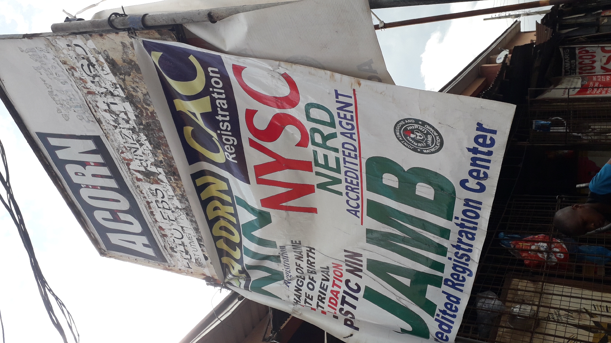 The image shows a faded sign with the word "ACORN" prominently displayed at the top. Below it, a large banner lists various services, including "NIN Registration," "NYSC NERD ACCREDITED AGENT," and "JAMB Accredited Registration Center." The banner appears to be made of fabric and is attached to a pole. The overall environment suggests a commercial area, with buildings and a storefront visible in the background. The image was taken in Onitsha, Nigeria.