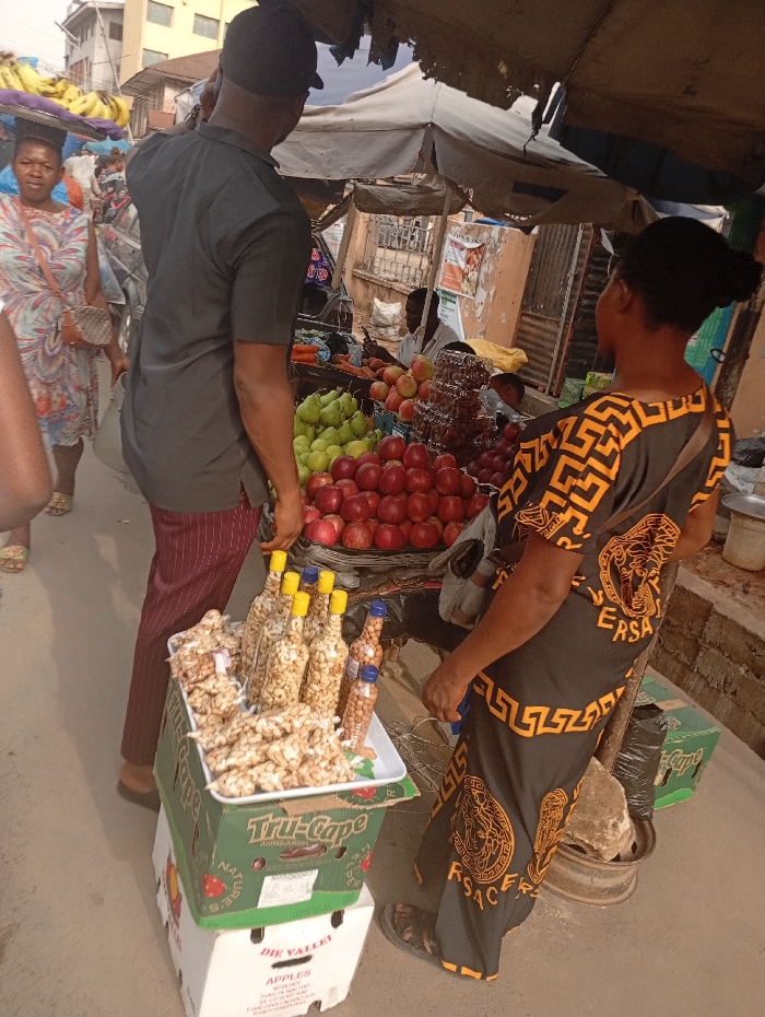 The image captures a vibrant outdoor market scene during the daytime, filled with people and fresh produce.
In the foreground, slightly to the left, a man is seen from behind, wearing a dark grey short-sleeved shirt, dark cap, and striped maroon and black trousers. He is looking towards a fruit stall. Next to him, on the right, a woman also stands with her back to the camera, dressed in a striking black and gold patterned long dress, featuring a distinctive Greek key motif and what appears to be the "VERSACE" logo repeating. She is wearing dark sandals and is also looking towards the fruit display.
Positioned in front of the man and woman, and slightly to the left, a white plastic tray holds a generous pile of what appear to be roasted peanuts or similar light-colored snacks. Several clear plastic bottles, mostly with yellow caps but one with a blue cap, are filled with peanuts or cashews and are also displayed on this tray. This tray rests on two stacked cardboard boxes. The top box is green and white with the visible text "Tru-Cape" and "APPLES", while the bottom box is white and shows "DIE VALLEY" and "APPLES".
The main feature of the scene is the market stall under a makeshift, somewhat tattered canopy. The stall is laden with a colorful array of fresh fruits. Prominently displayed are piles of green pears and bright red apples. Further back, red grapes are visible in clear plastic containers, and there might be other red fruits like pomegranates. Some vegetables, possibly carrots, can be glimpsed in the background. Behind the stall, at least one person, likely a vendor, is partially visible.
In the mid-ground on the left, another woman is walking by, carrying a large basket of bananas on her head, a common sight in such markets. She is wearing a patterned dress.
The background shows other market activity, faint posters on a wall, and parts of urban buildings under a clear sky, suggesting a busy street or market square. The overall impression is one of a bustling local market with various goods on offer.