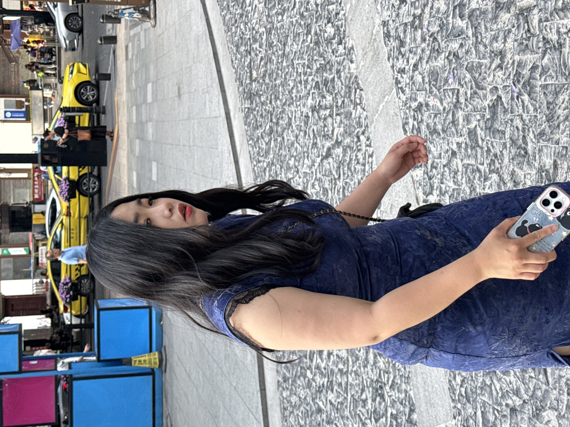 A medium shot shows a woman with long, dark hair, wearing a blue dress and holding a cell phone with a cartoon design. She is standing on a sidewalk in Yuzhong District, China. To the left of the woman, there are taxis and various blue and pink structures, possibly booths or displays. The sidewalk is gray and appears to be made of stone or textured concrete.