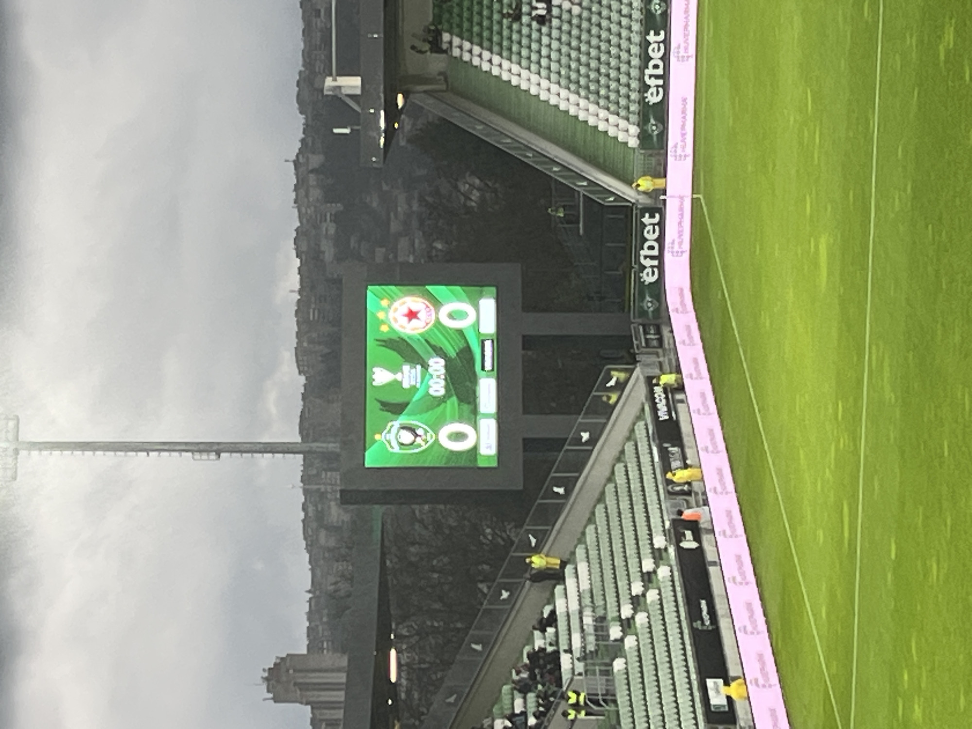 The image depicts a football stadium in Razgrad, Bulgaria, captured on an overcast day. The main focus is a large video scoreboard displaying details for a football match. The scoreboard shows the logos of two teams: CSKA Sofia (represented by a red star) and PFC Ludogorets (represented by a green eagle). Both teams have a score of "0," and the match clock reads "00:00," indicating the very beginning of a game, a pre-match setup, or possibly halftime. A trophy icon is also visible on the scoreboard, suggesting a cup competition.
The stadium features rows of green and white seats, which appear mostly empty, with only a few people, likely staff or security personnel in high-visibility vests, scattered throughout the stands and near the pitch. The lush green football field is visible on the right, bordered by digital advertising hoardings. These hoardings prominently display the brand name "efbet" multiple times, along with "VIVACO" and "БУЛВЕРИА".
In the background, beyond the stadium's upper structures, a cloudy sky dominates, with distant buildings and a cityscape visible beneath it. A tall stadium floodlight tower extends upwards on the left side of the frame. The scene suggests a quiet moment at the stadium, possibly before spectators arrive for a match or during an event with limited attendance.