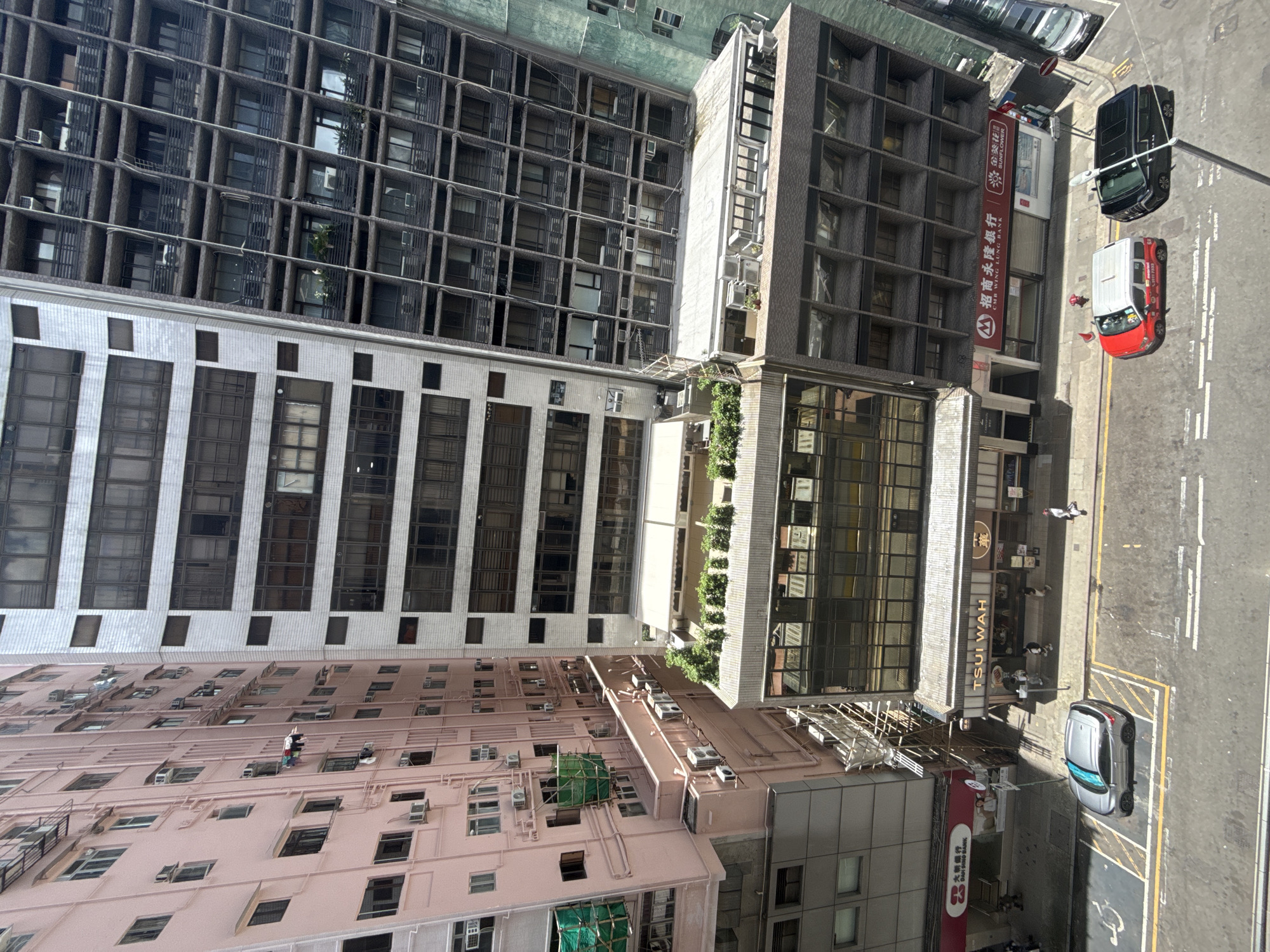 The image captures a street scene in Hong Kong, China, featuring a row of tall buildings. The buildings are a mix of architectural styles and colors, with the pink building on the left appearing to be residential, characterized by numerous windows and external pipes. Adjacent to it is a white building with a grid-like facade, followed by a darker, more modern building with balconies and multiple windows. The ground level of these buildings houses various businesses, including a bank with a prominent red signage and a restaurant identified by the "TSUI WAH" sign.
The street below is paved with asphalt and marked with yellow and white lines. Several cars are visible: a silver SUV is parked in a designated spot, a red and white vehicle, possibly a taxi or van, is stopped, and a black vehicle is parked further down the street. Pedestrians are sparsely distributed on the sidewalks, some walking and others seemingly interacting with the establishments. Scaffolding is visible on one of the buildings, suggesting ongoing construction or maintenance. The overall impression is of a busy urban environment with a dense concentration of commercial and residential structures typical of Hong Kong.