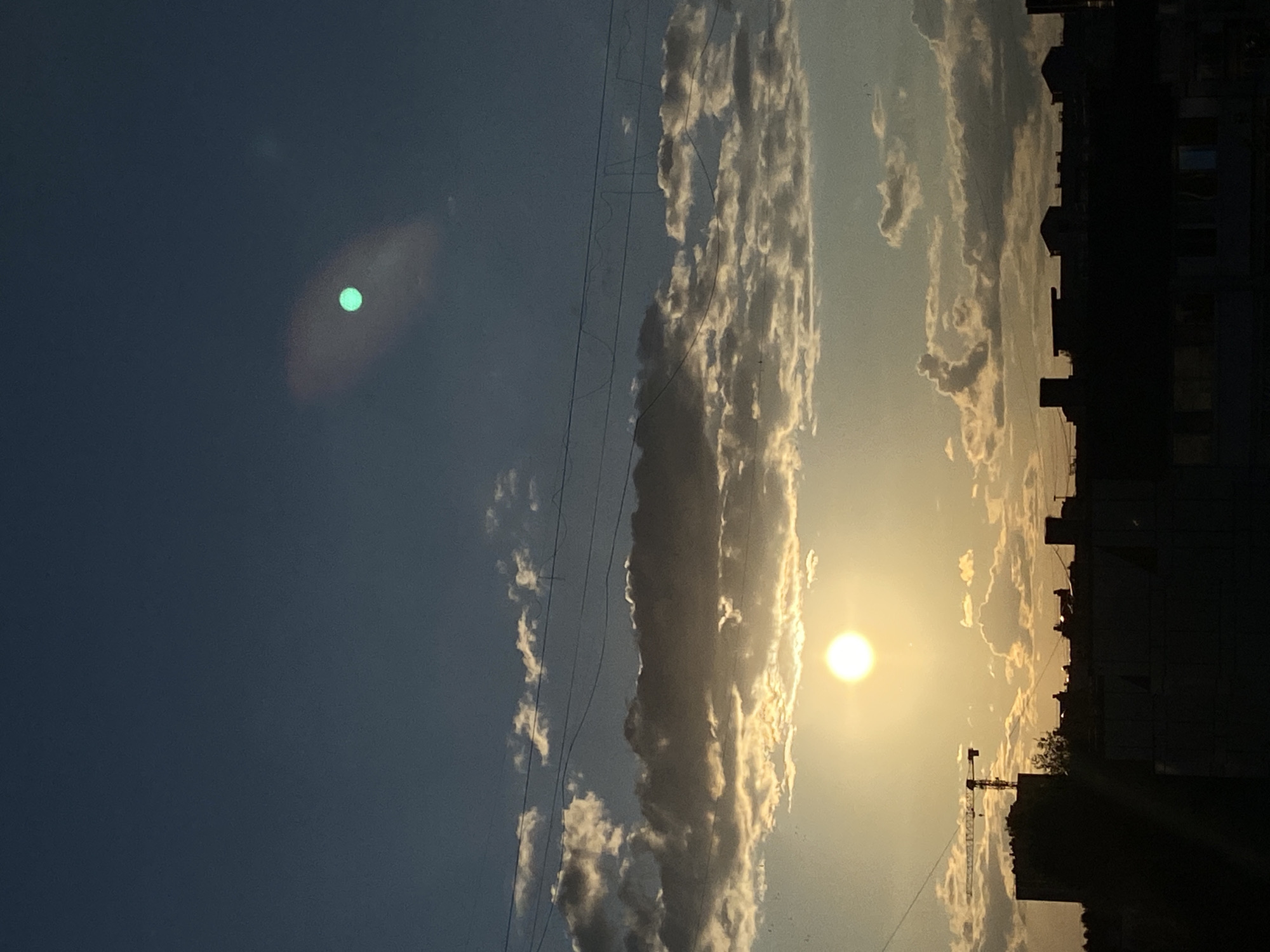 A silhouette of the city of Chaikovsky, Russia, is captured against a sunset sky. The foreground is dominated by the dark shapes of buildings and a construction crane. The sky is a gradient of deep blue at the top, transitioning to lighter blues and yellows near the horizon, illuminated by the bright, setting sun. Clouds are scattered throughout the sky, some catching the warm light of the sunset and appearing golden, while others are darker and more shadowed. In the upper right portion of the sky, a bright, turquoise-colored circular object is visible, possibly a lens flare or a light artifact. Power lines are visible horizontally across the sky, partially obscuring the clouds.
