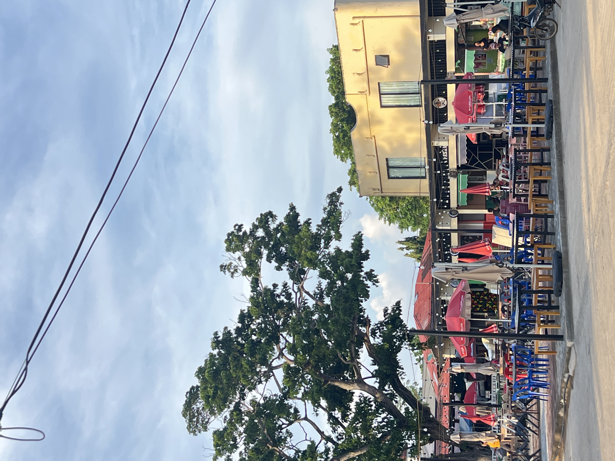 The image depicts an outdoor seating area at a restaurant or cafe in Cuyo, Philippines. The scene is set on a paved surface, likely a street or plaza, with an overcast sky overhead.
In the foreground, numerous tables and chairs are arranged in a scattered fashion, suggesting a casual dining environment. Many of the tables are occupied by chairs of various colors, including blue, red, and white. Several red umbrellas, some open and some closed, are positioned over the tables, providing shade.
A large, leafy tree dominates the left side of the frame, its branches extending over a portion of the seating area. Beyond the tables, a building with a yellow facade and dark-framed windows is visible. A covered walkway or patio runs along the front of this building, and more tables and chairs are set up beneath it. String lights are suspended along this walkway.
In the background, additional structures with red roofs are present. Some people are visible in the distance, including a person walking near the tree on the left and individuals seated at tables in front of the yellow building. A motorcycle is parked on the right side of the image. The overall impression is a vibrant, communal outdoor space, typical of a local establishment in the Philippines.