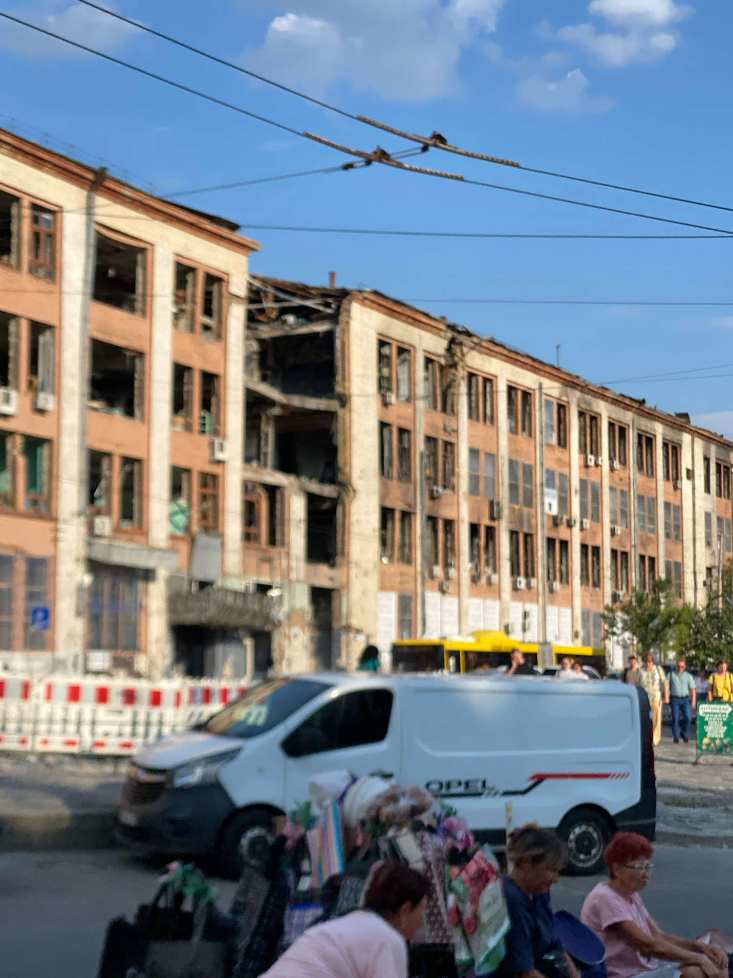 An eye-level, slightly blurry, long shot captures a street scene in front of a damaged, multi-story building under a clear blue sky with scattered clouds. The building, appearing to be an apartment or office complex, shows significant damage with many windows broken and sections of the roof and facade destroyed, revealing structural damage. The building's exterior is a light beige with darker, orange-brown trim around the windows and roofline. Several air conditioning units are visible on the facade.
In the foreground, a white Opel van is parked on the street. In front of the van, to the left, are some carts filled with goods, and to the right, three women are seated on chairs, facing away from the camera, with one woman looking towards the camera. A yellow bus is partially visible behind the van. Further in the background, pedestrians are walking along the sidewalk. Construction barriers with red and white markings are placed along the street in front of the building. Power lines are visible across the top of the frame.