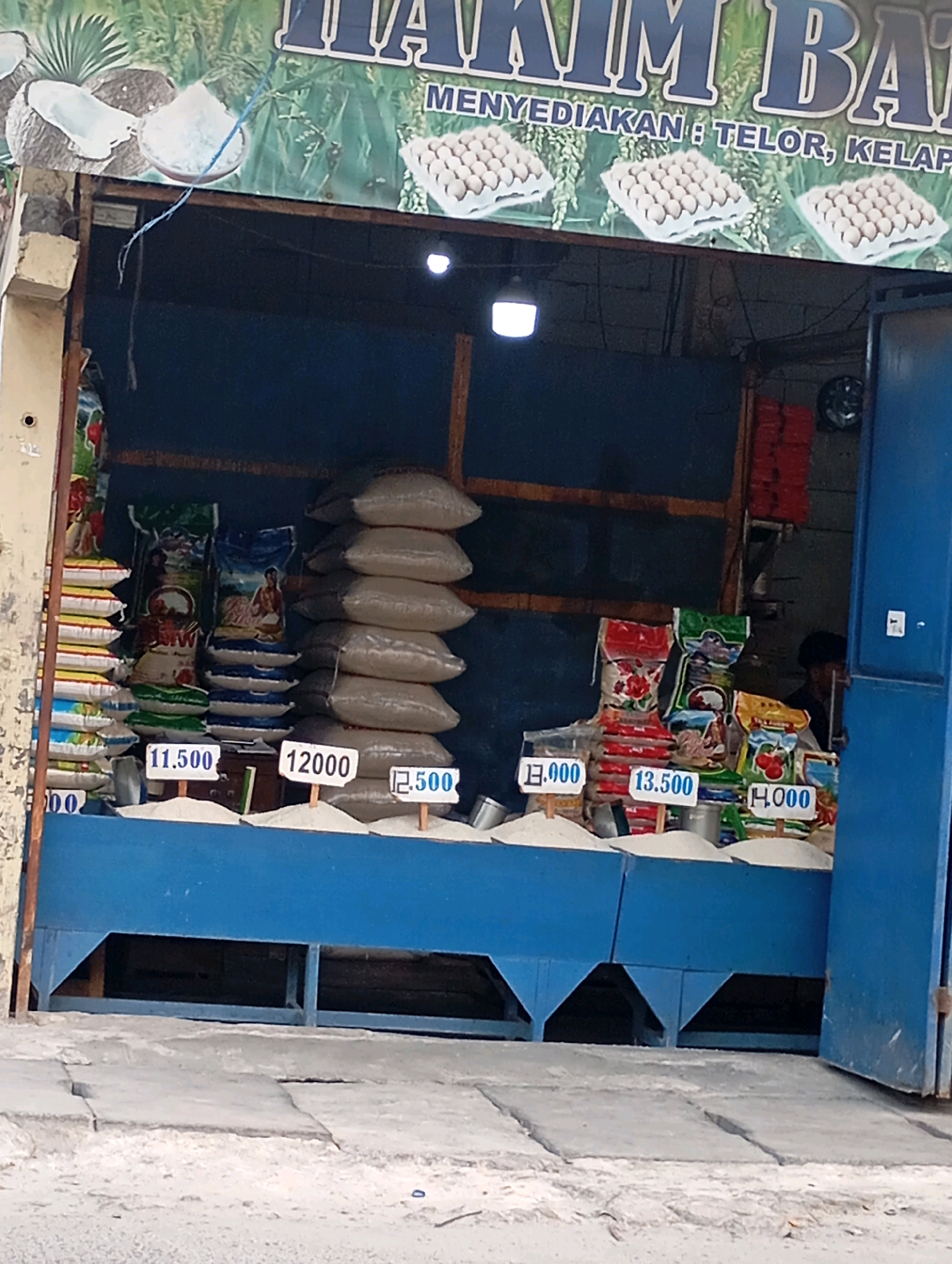 The image depicts a storefront in Babelan, Indonesia, likely a small grocery or market stall. The facade features a blue overhang and a blue counter where various sacks of rice and bags of other goods are displayed. Prices are visible on small white signs placed in front of the rice sacks, indicating prices such as "11.500", "12000", "12.500", "13.000", "13.500", and "H.000". Stacks of colorful bags, possibly containing rice or other dry goods, are arranged on the left side of the stall. In the background, a person with dark hair is partially visible behind the counter. Above the stall, a banner displays text in large blue letters, partially obscured, but it appears to advertise products such as eggs and coconuts, with text that includes "MENYEDIAKAN : TELOR, KELAP...". The ground in front of the stall is paved with concrete slabs, with some loose sand or dust present. The overall scene suggests a local market or small business in an urban or semi-urban environment in Babelan.