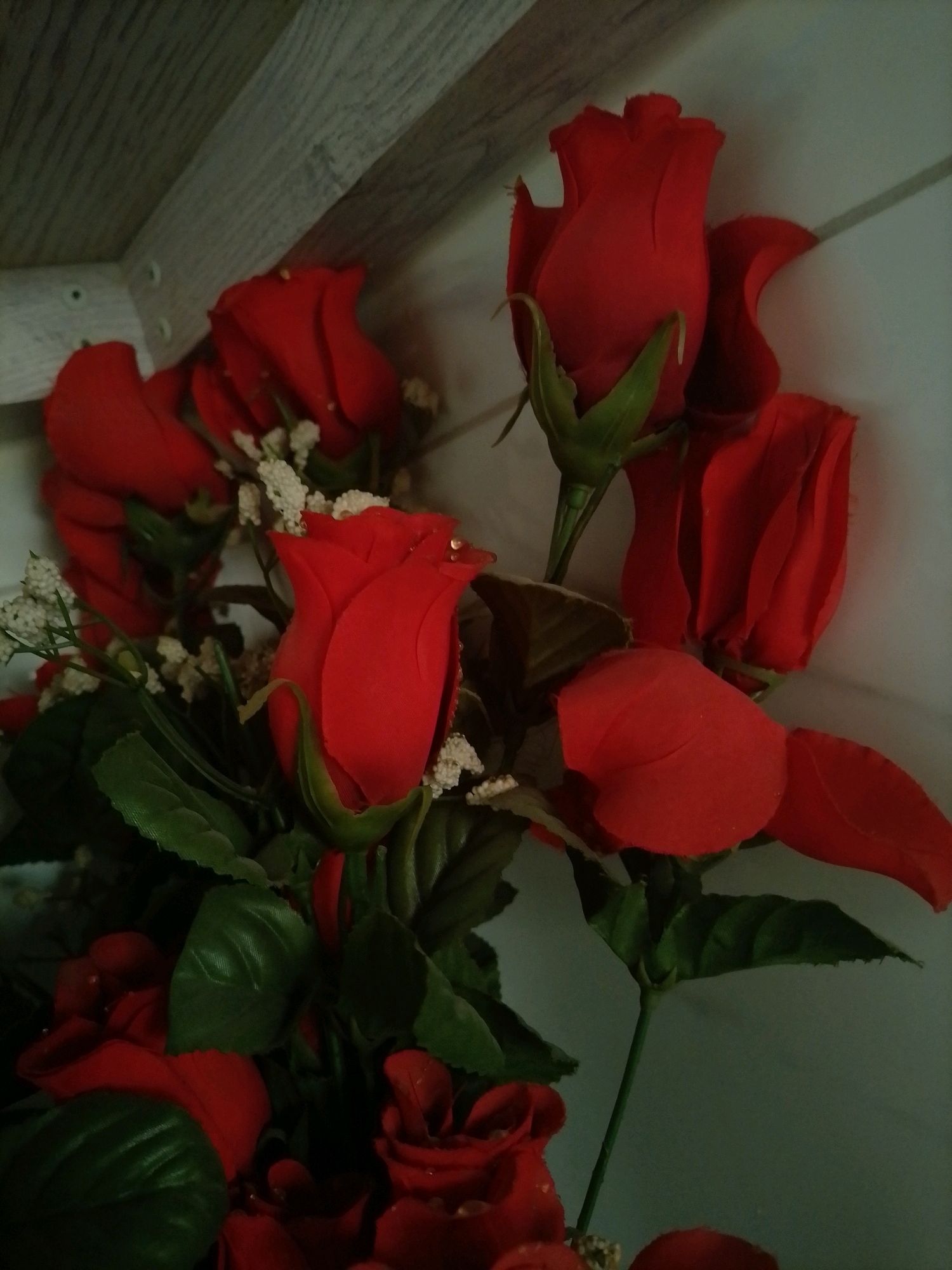 A close-up, slightly overhead shot shows a bouquet of artificial red roses with green leaves and small white filler flowers. The flowers are arranged in a loose cluster. The background is a soft, off-white wall with faint vertical lines, and a section of a light-colored wooden surface with visible grain and some screws is visible in the upper left corner. The lighting is dim, casting shadows on the subjects. There are no people or visible signs of life. The image's composition suggests it was taken indoors, possibly on a shelf or table. The specific location in Amman, Jordan, cannot be determined from the visual information provided.