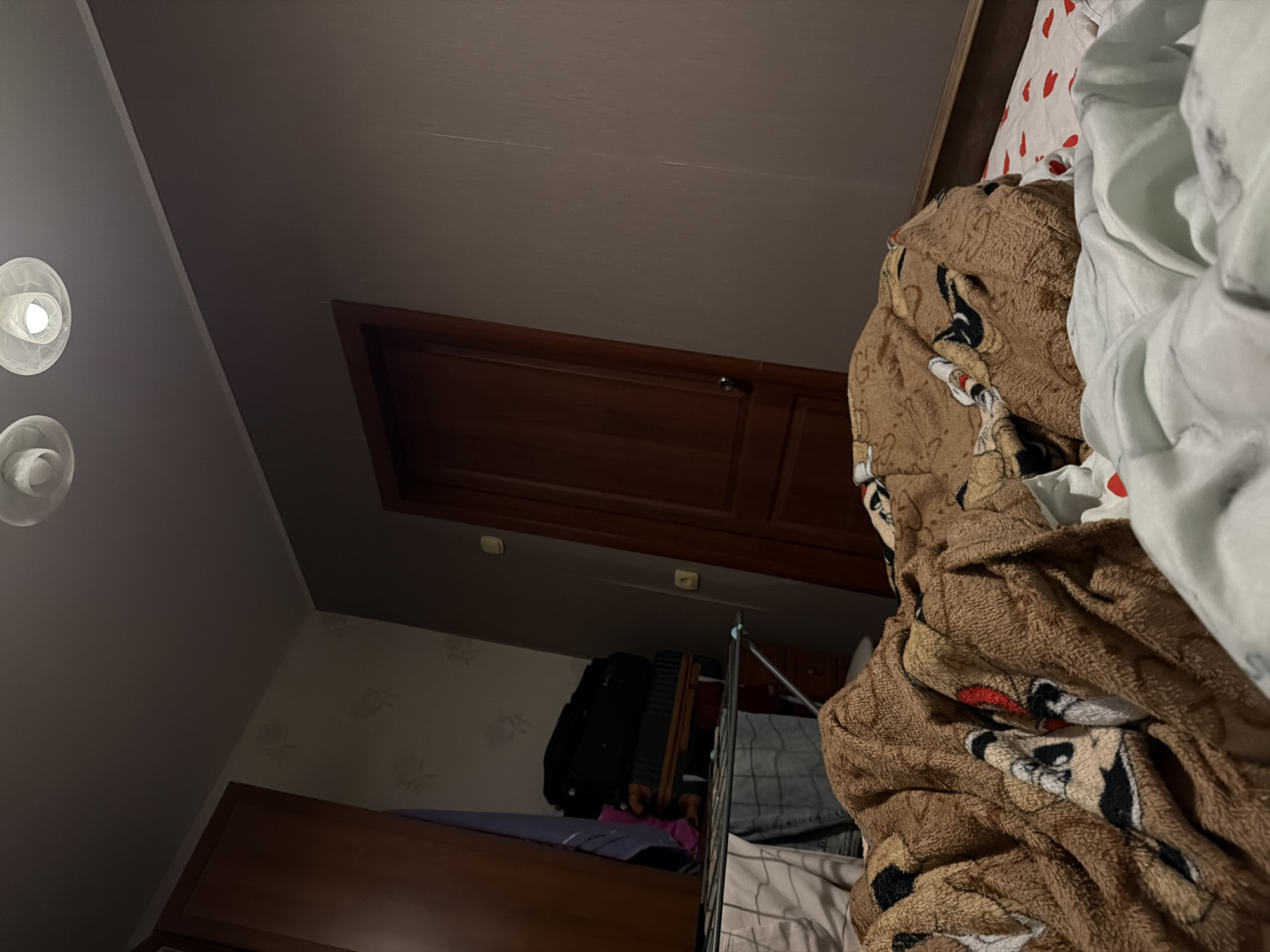 An indoor, low-light scene depicts a bedroom with a bed in the foreground, covered by a brown blanket with cartoon characters. A white duvet is partially visible in front of the blanket. To the left of the bed, a drying rack holds various items of clothing, and a stack of luggage is placed against a wooden wardrobe. Further back, a closed wooden door stands against a wall with textured wallpaper. The ceiling features two circular light fixtures, one of which is illuminated. The overall impression is one of a lived-in, slightly cluttered personal space.
