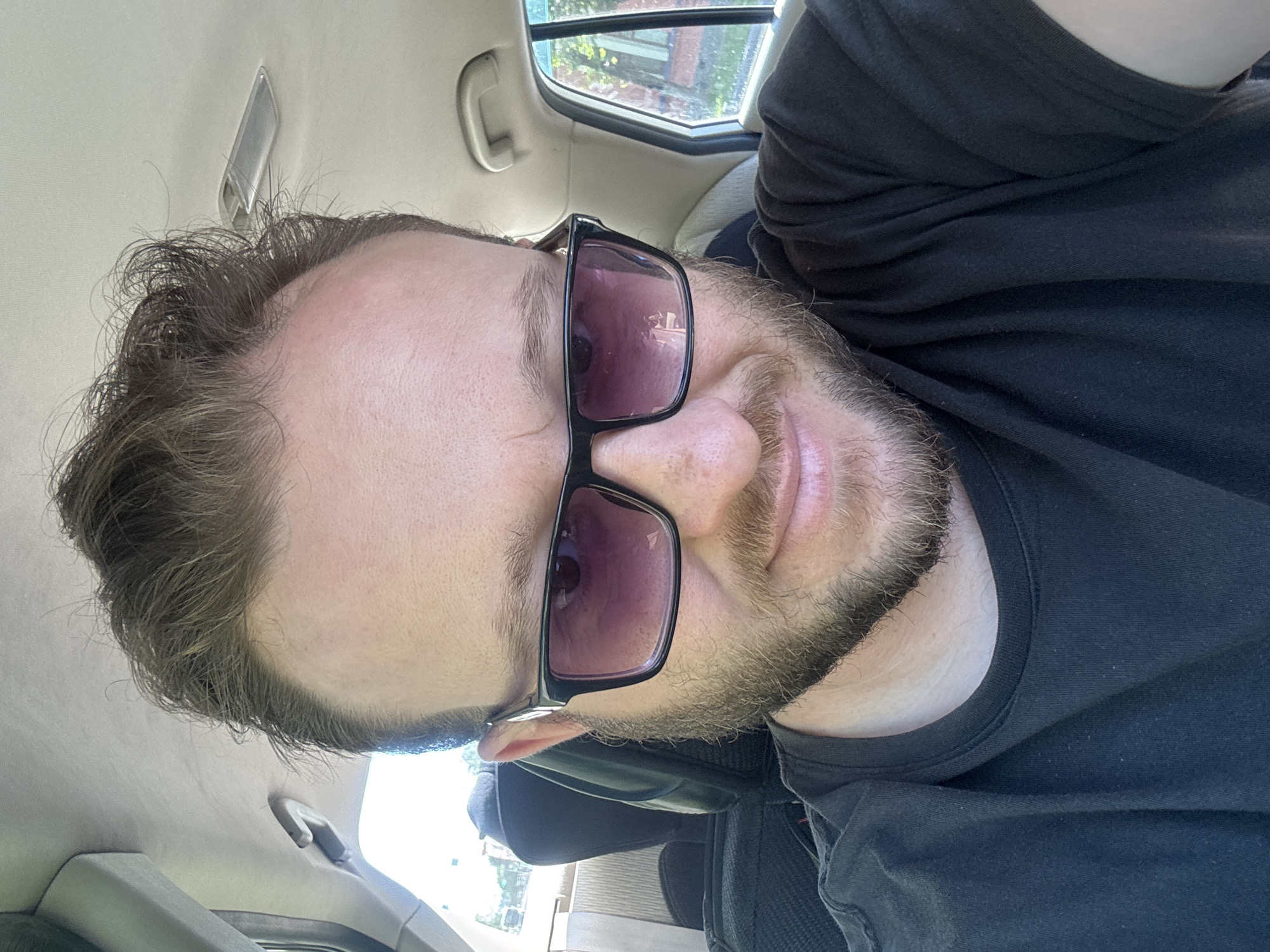 A man wearing sunglasses and a black t-shirt is sitting in the passenger seat of a car. He has brown hair and a beard. The interior of the car is visible, including the headliner, a door handle, and the back of a seat. A window in the car shows trees and buildings outside. The image was taken during the day. There is no information about the location being Blagoveshchensk, Russia.