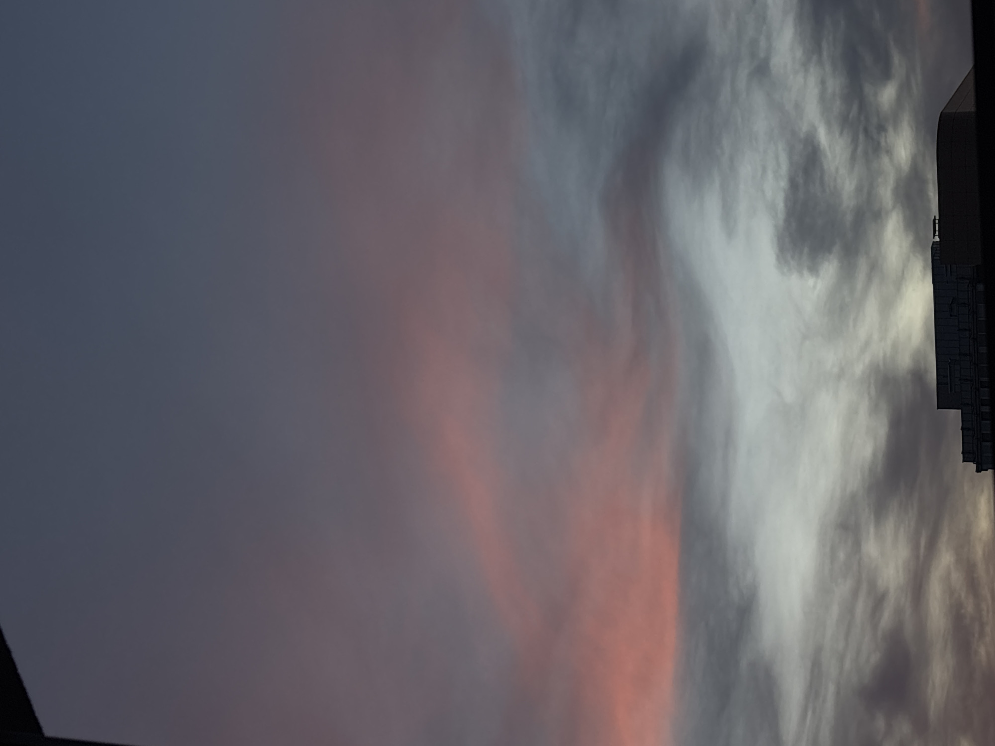 The image shows a view of the sky in Belgrade, Serbia, with clouds tinged with pink hues from the setting sun. A dark, indistinct building is visible on the right edge of the frame. The sky transitions from a darker, overcast gray in the top left to lighter gray and pinkish tones toward the right, indicating a sunset or twilight scene.
