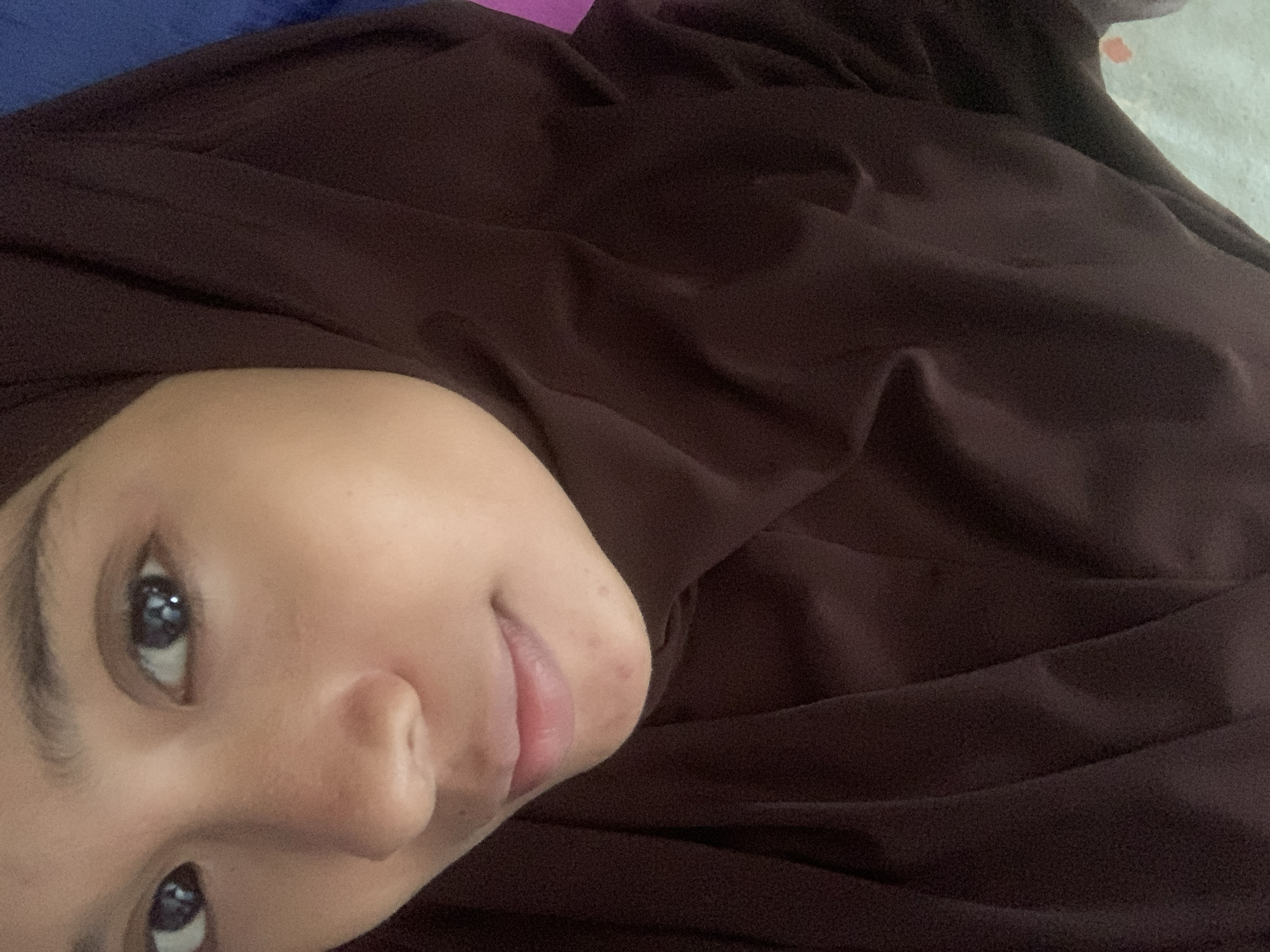 A close-up, self-portrait image depicts a young woman's face, partially covered by a dark brown hijab. The focus is on her eyes, nose, and mouth. Her dark eyes are looking directly at the camera. The skin on her face appears to have some minor blemishes. The background is blurred, with a hint of blue and pink on the right side, suggesting an indoor setting. The image was taken in Sungai Meriam, Indonesia.