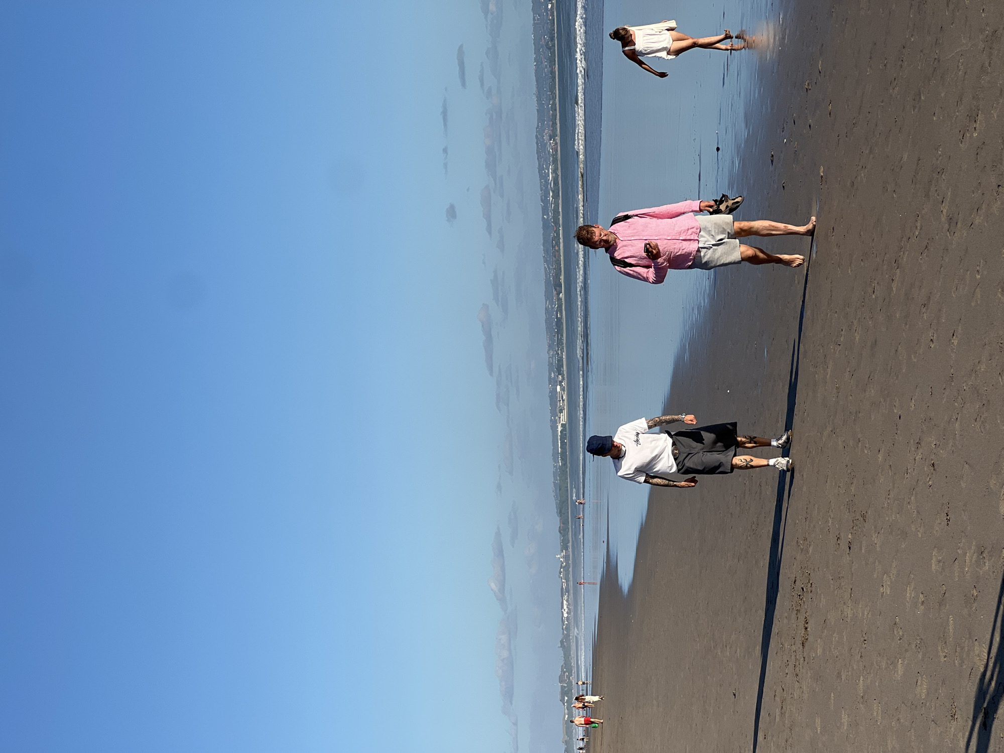 This image depicts a view of Kuta Beach in Kuta, Indonesia, under a clear blue sky with scattered clouds. The sandy beach stretches into the distance, with gentle waves lapping at the shore. In the foreground, three individuals are walking along the wet sand. A man with tattoos on his arms and legs, wearing a white t-shirt and dark shorts, walks towards the camera. To his right, another man in a bright pink shirt and grey shorts is looking at his phone while carrying a pair of shoes in his hand. Further in the distance, a woman in a white dress is walking away from the camera. Other beachgoers can be seen scattered along the shoreline and in the shallow water, with buildings and the coastline visible in the background. The lighting suggests it is late afternoon or early morning.