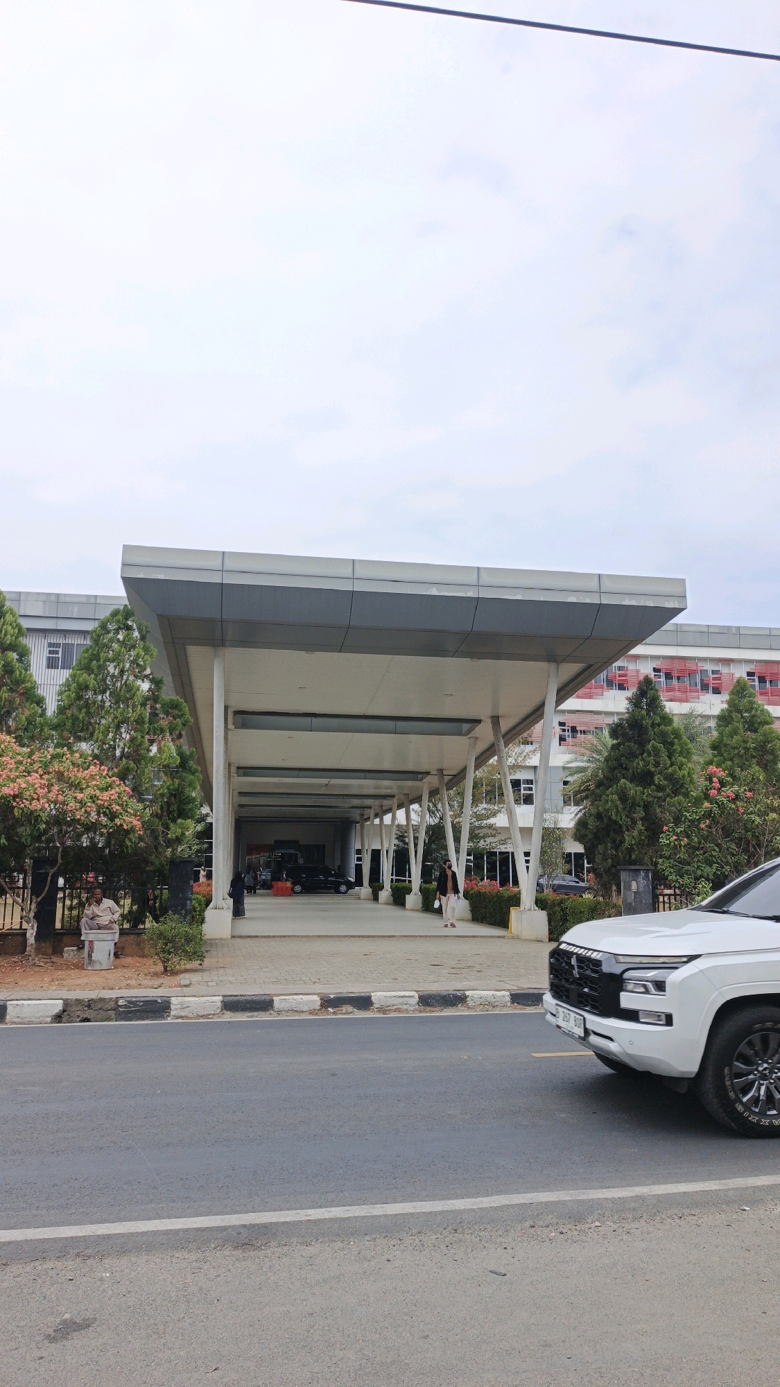 The image captures a daytime exterior view of a modern building with a prominent covered entryway. The building appears to be a hospital or a large institutional facility, with multiple stories and a red and white facade visible in the background. The covered entryway is supported by white, angular columns and has a flat, grey roof. Several cars are parked or driving under the entryway, and a few pedestrians are visible walking.
In the foreground, a white SUV is parked on the right side of the frame, facing towards the building. The road in front of the building is paved and has a sidewalk with a black and white curb. Trees and some flowering bushes are planted along the sidewalk. A man is sitting on a concrete block on the left side of the sidewalk, looking towards the building.
The sky is overcast and a dark wire is visible stretching across the top of the image. The overall scene suggests a functional, public space in an urban or semi-urban setting. The license plate on the white SUV appears to have Indonesian characters, and the building's architecture is consistent with modern construction found in Indonesia. Based on the license plate and architectural style, this image could be from Pangandaran, Indonesia.