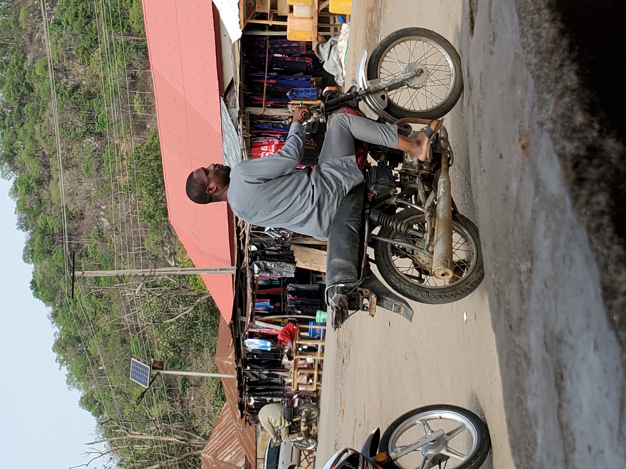 The image captures a street or market scene in Mararaba, Nigeria, during daylight hours under a clear sky. The main subject is a man with dark skin, short hair, and a beard, wearing a light grey long-sleeved tunic and dark grey trousers, along with sandals. He is seated on a black and silver motorcycle, appearing stationary, with his hands near the handlebars, looking slightly to his left.
Behind the man, market stalls with red corrugated metal roofs line the roadside, displaying various items, including clothing and what appears to be general goods. Another person is partially visible further back within one of the stalls. The road surface is visible in the foreground, appearing somewhat rough or dusty.
In the background, a large, tree-covered hill or mountain dominates the landscape. Utility poles with multiple power lines extend across the upper left portion of the image, and one pole features a small solar panel attached. Parts of a second motorcycle, including a wheel with "Haolue" faintly visible on the tire, are seen in the lower right foreground. The overall impression is that of a bustling local area.