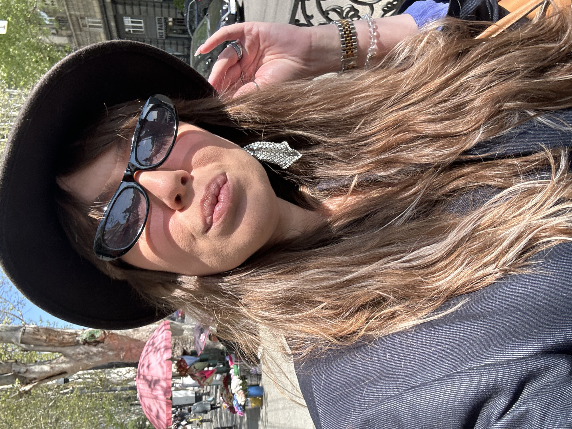 A close-up, tilted portrait captures a woman outdoors on a bright, sunny day, likely in an urban setting such as Tbilisi, Georgia. The woman is the primary subject, positioned diagonally across the frame. She wears a dark brown or black wide-brimmed hat, black sunglasses, and a dark blue or grey pinstriped jacket. Her long, wavy hair, a mix of light brown and blonde hues, cascades across her shoulder and chest. A sparkling, silver-toned hair clip is visible in her hair above her right ear.
Her left hand is raised, with manicured nails, revealing a silver ring, a gold and silver watch, and a clear beaded bracelet on her wrist. The bright sunlight creates strong highlights and shadows, particularly on her face and hair. Her lips are slightly parted.
In the background, an old multi-story building with windows is visible, and faint text, possibly "PIZZA BAR," can be discerned on its facade. To the left and below the woman's head, parts of a large tree trunk are seen, along with a partial view of a pink umbrella and what appear to be street market stalls or vendors' goods, suggesting a lively outdoor environment. The time of day is clearly daytime, with clear, sunny weather.