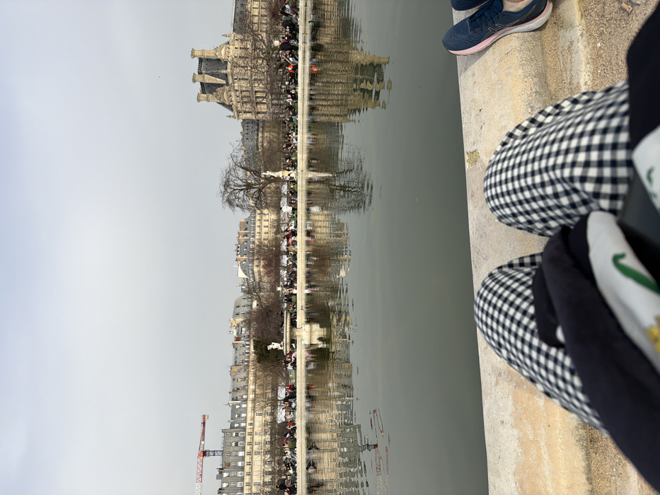 The image depicts a scene from the Jardin des Tuileries in Paris, France, specifically from the edge of the Grand Bassin Rond. The perspective is low-angle, from the viewpoint of a seated individual whose lower legs, clad in black and white gingham pants, occupy the immediate foreground. A dark garment rests on the lap, partially obscuring a white garment with green and gold detailing. To the right, a pair of dark blue and pink athletic shoes are positioned on the light-colored concrete coping of the basin.
The midground consists of the still, dark grey-green water of the large circular basin, reflecting background structures and bare trees. Beyond the basin, numerous individuals are visible, seated on green park chairs or standing, populating the park grounds along the distant edge. The background skyline features a continuous line of classic Parisian Haussmannian buildings with grey zinc roofs on the left and center, above which a red and white construction crane is prominently visible. Interspersed among bare deciduous trees are white statues and a central fountain. On the far right, a large, ornate building with multiple distinct chimneys and a dark mansard roof, consistent with the architecture of the Louvre Palace, stands against the uniformly overcast light grey sky.