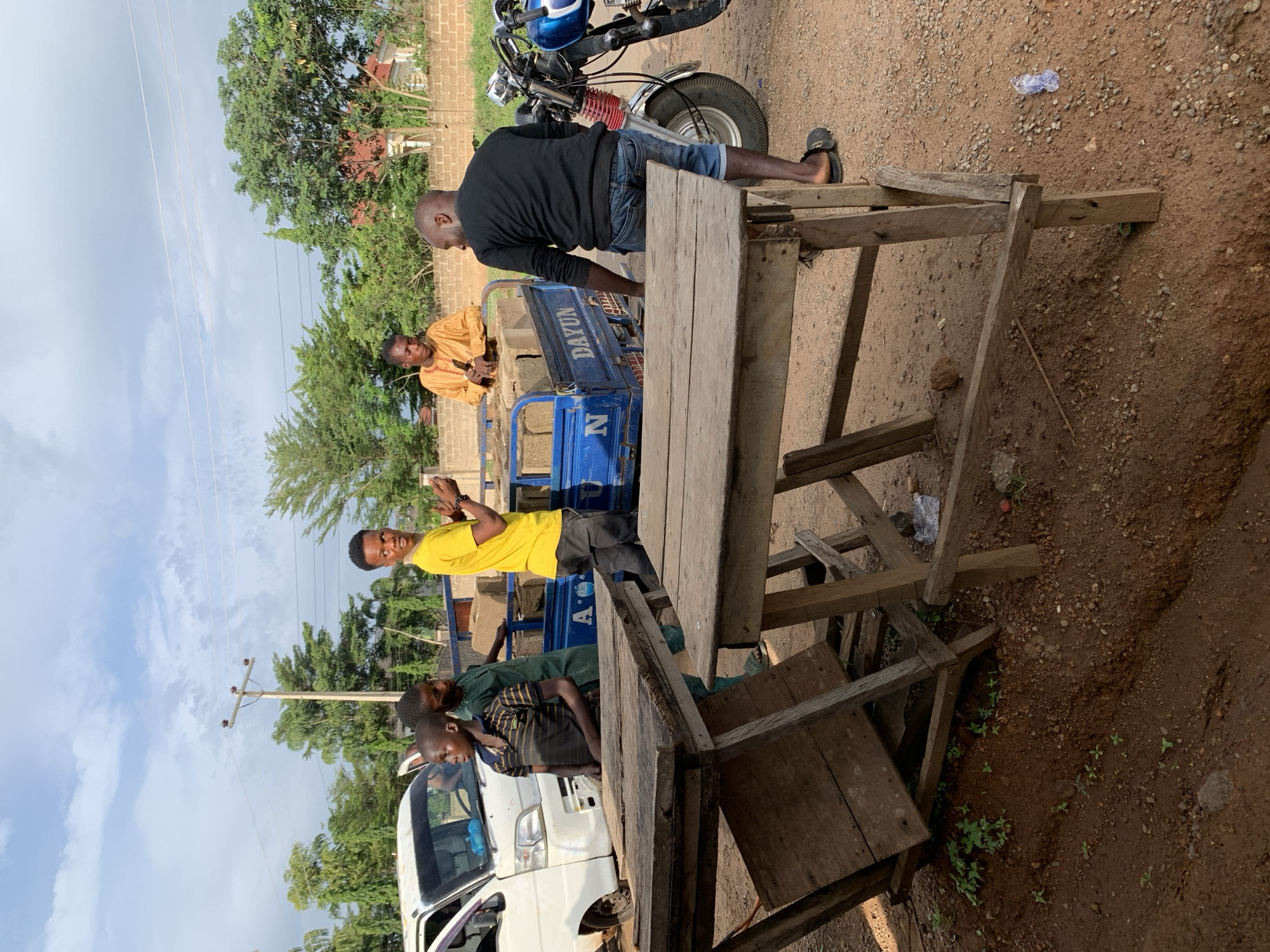 The image shows a street scene in Kabba, Nigeria. A blue tricycle with the word "DAYUN" on the side is partially visible, loaded with rectangular objects that appear to be concrete blocks. A man in a long-sleeved orange shirt sits on top of the load. Another man, wearing a black long-sleeved shirt and blue jeans shorts, stands at the back of the tricycle, seemingly observing. Three young boys are near a white van, one in a yellow shirt holding up his hands, another wearing a green shirt leaning over the side, and a third wearing a striped shirt. In the foreground stands a rough, wooden table or counter made of unfinished planks. A motorcycle is partially visible at the top of the image. The ground is dirt. Trees and buildings are visible in the background.
