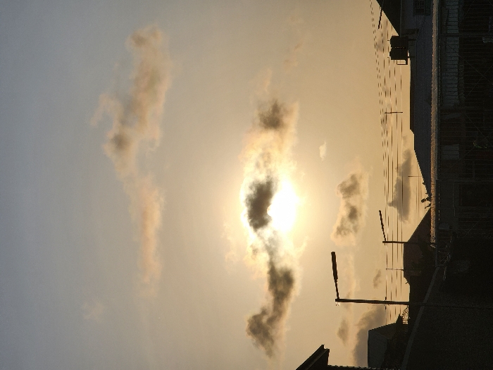 This image captures an urban landscape in Lekki, Nigeria, at either sunrise or sunset. The sun is centrally positioned in the upper mid-ground, partially obscured by dark, patchy clouds, radiating a strong light that transitions the sky from bright white-yellow around the sun to a pale blue-grey towards the top. Several smaller, diffused clouds are scattered across the sky.
The lower third of the image features a dark, jagged silhouette of multiple building rooftops, indicating a developed area. Two distinct streetlights or utility poles with horizontal fixtures are prominently silhouetted against the bright sky, one larger on the left and a smaller one further right. Numerous parallel utility wires extend horizontally across the mid-ground, connecting unseen poles. On one of the distant rooftops, two dark, cylindrical objects, likely water tanks, are visible. The immediate foreground is dominated by a dark, solid fence or wall, obscuring any ground-level details. No individuals or specific actions are discernible within the frame.