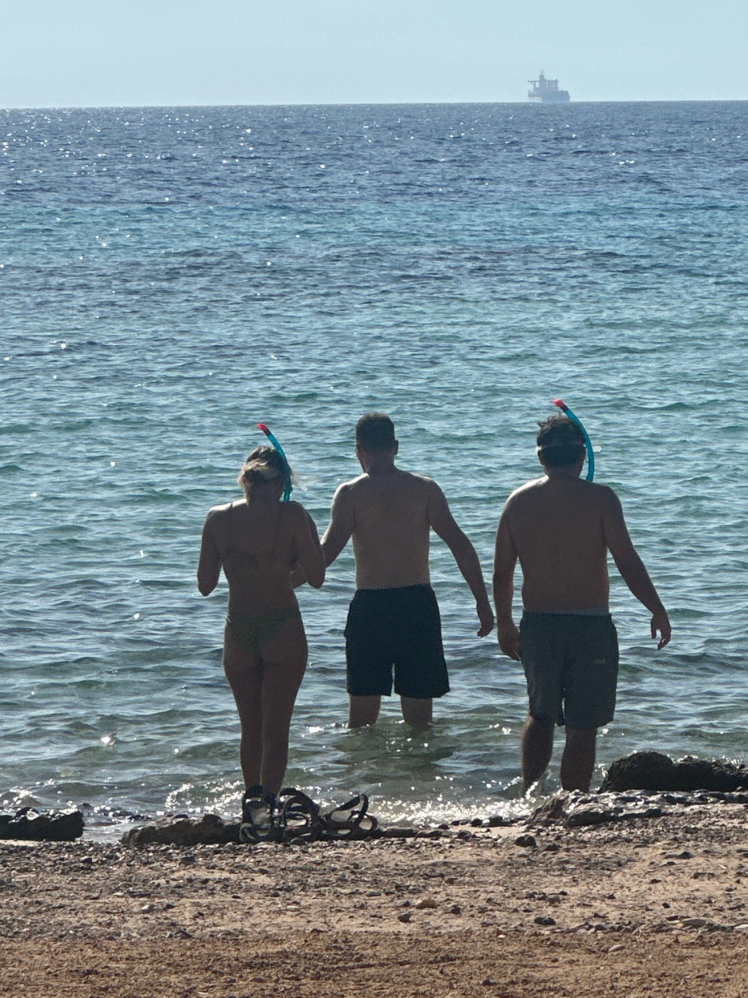 In this image, taken on the shores of Nuweiba, Egypt, three individuals are partially submerged in the clear turquoise waters of the Red Sea. The people, viewed from behind, are wearing snorkeling gear. The leftmost individual, a woman, is in a bikini. To her right are two men, one in dark shorts and the other in khaki shorts. The water's surface is rippled, reflecting the bright sunlight. In the distance, a large cargo ship is visible on the horizon, appearing as a dark silhouette against a hazy sky. The foreground consists of a rocky, sandy beach with a few scattered pieces of driftwood and a pair of sandals. The overall scene depicts a serene beach setting with people preparing to snorkel.
