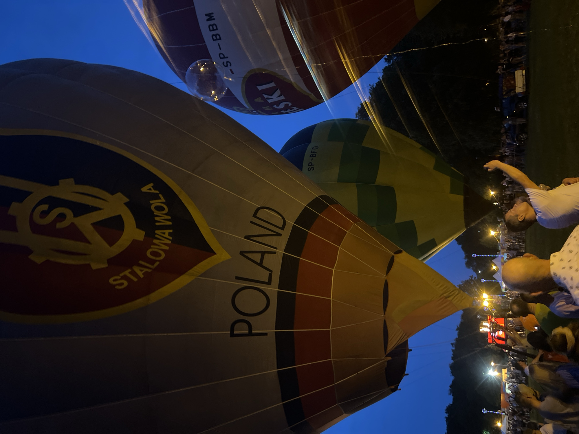 This low-angle, close-up shot captures a crowd gathered at a hot air balloon festival at night, likely in Tychy, Poland, given the "POLAND" inscription on one of the balloons. The foreground is dominated by the illuminated, partially inflated bodies of several hot air balloons. The largest balloon in the foreground has "POLAND" written across it in bold black letters, with bands of red and black. Above this, a crest with the letters "S" and "A" within a gear shape, along with the text "STALOWA WOLA," is visible on a red and yellow section. To the right, another balloon displays "SP-BBM" and "MA-ESKI". A third balloon, green and yellow, is visible between the two main balloons, showing "SP-BFO". The sky is dark blue, suggesting it is late evening or night. In the background, a crowd of people is visible, with numerous lights from tents, lampposts, and possibly the burners of the hot air balloons creating streaks of light across the image. Some individuals in the crowd are holding up phones, likely capturing the event. The atmosphere appears festive.